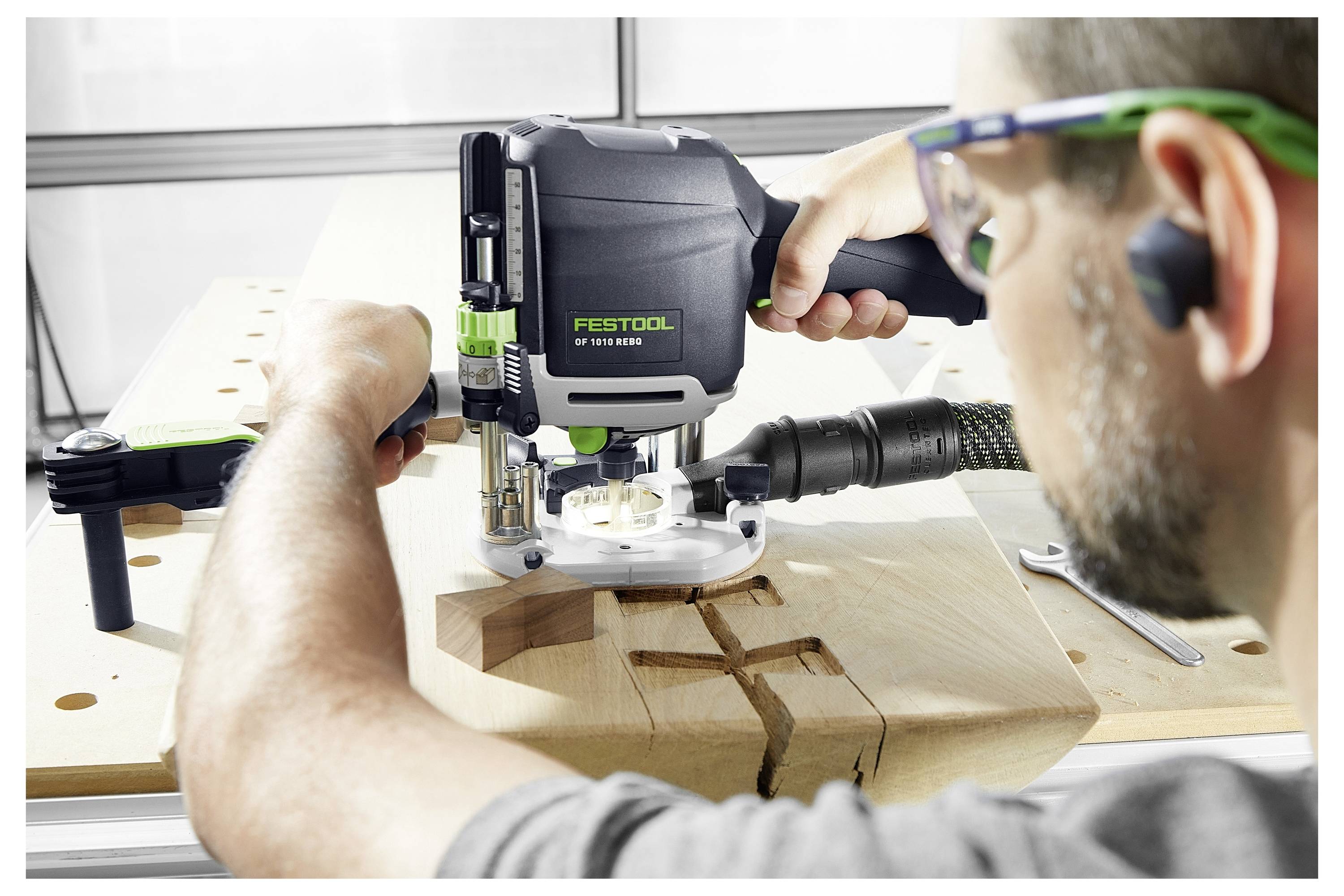 Eine Person nutzt einen Festool-Fräser, um ein präzises Muster in eine Holzplatte zu fräsen, wobei der Fokus auf aufwändiger Holzbearbeitung mit Schutzausrüstung liegt.