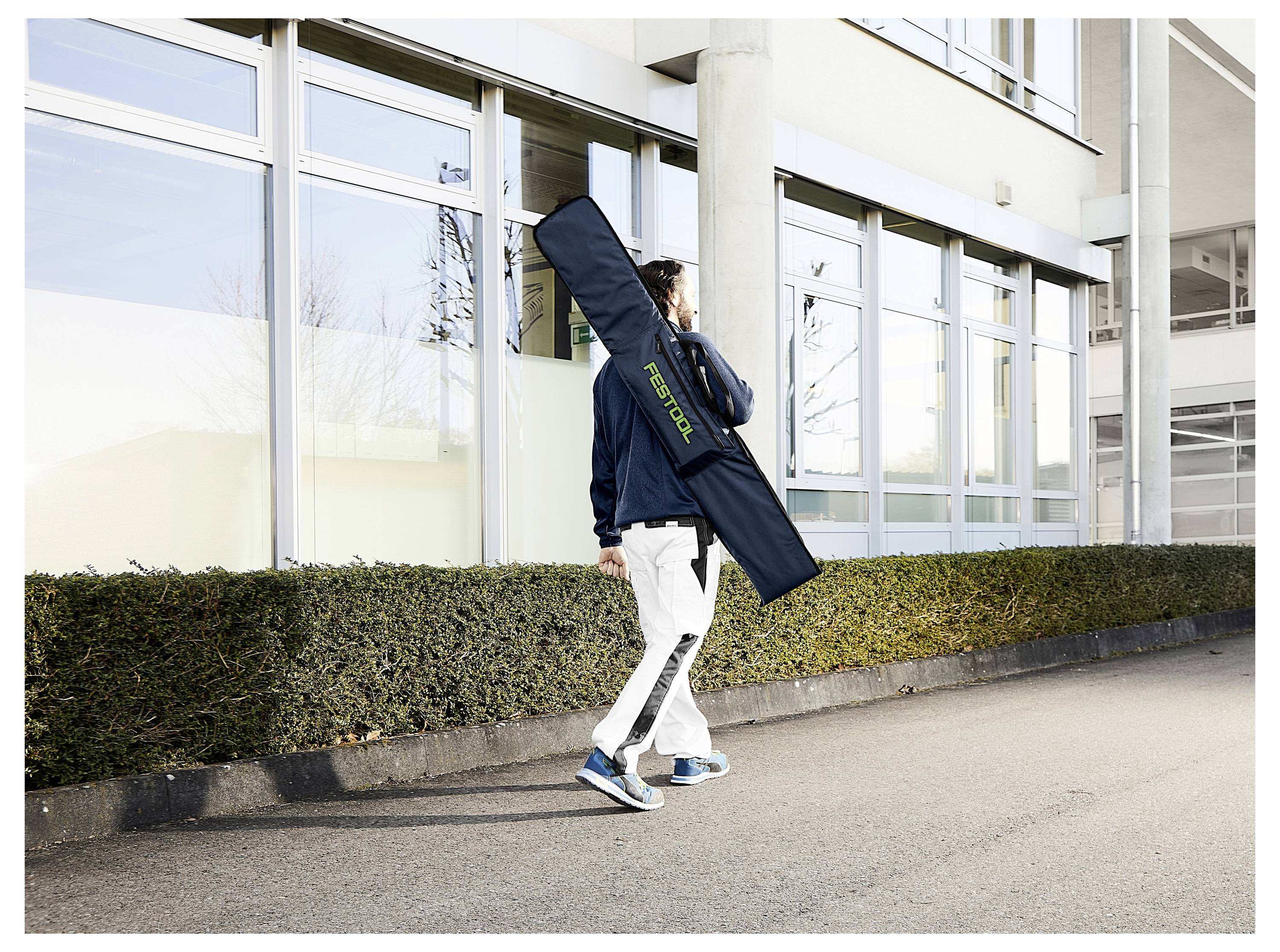 Eine Person, die auf einem Gehweg neben einem Gebäude spaziert und einen langen, schwarzen Koffer mit der Aufschrift 