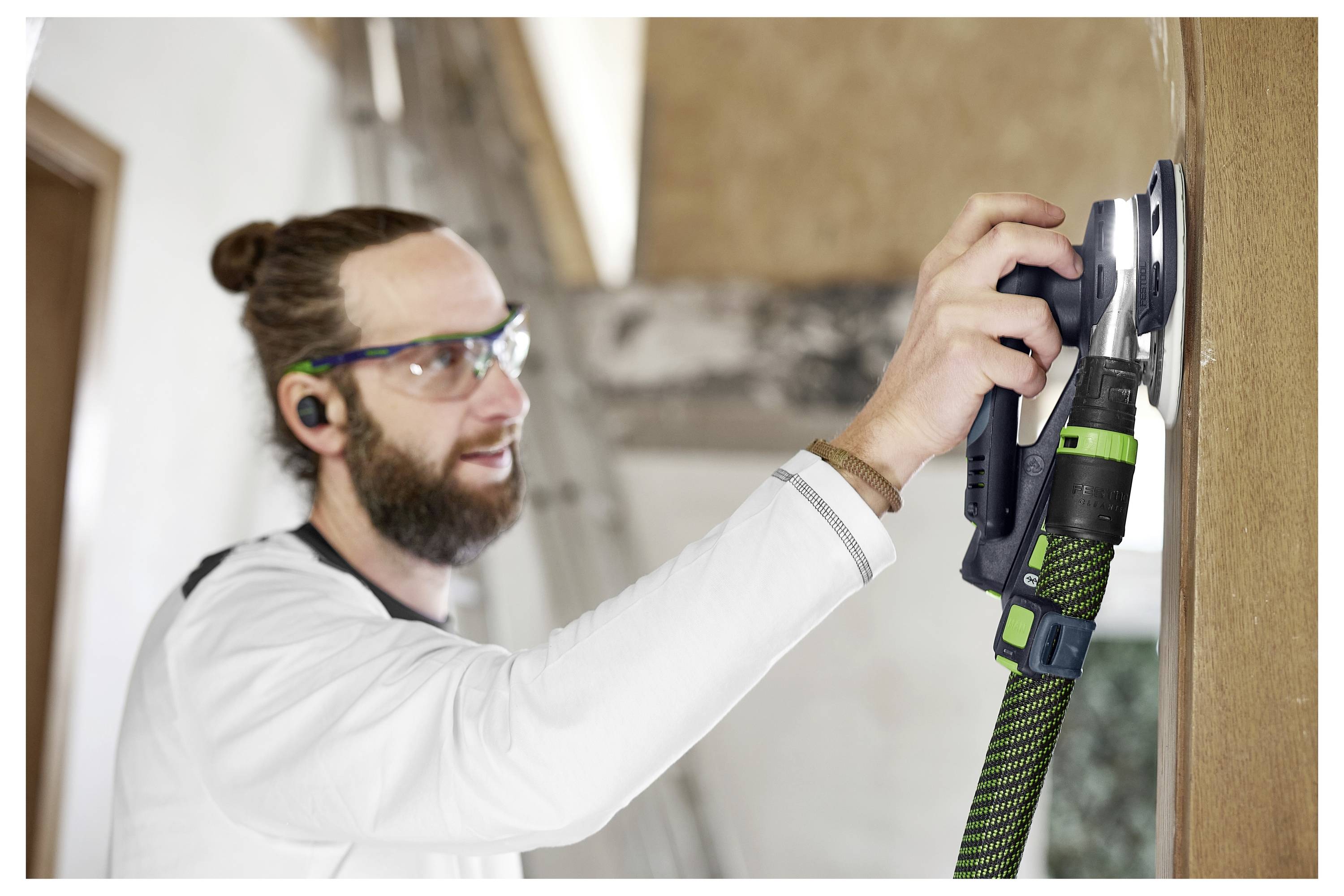 Eine Person, die eine Schutzbrille und einen Gehörschutz trägt, verwendet einen Elektroschleifera auf einer Holzoberfläche in Innenräumen.