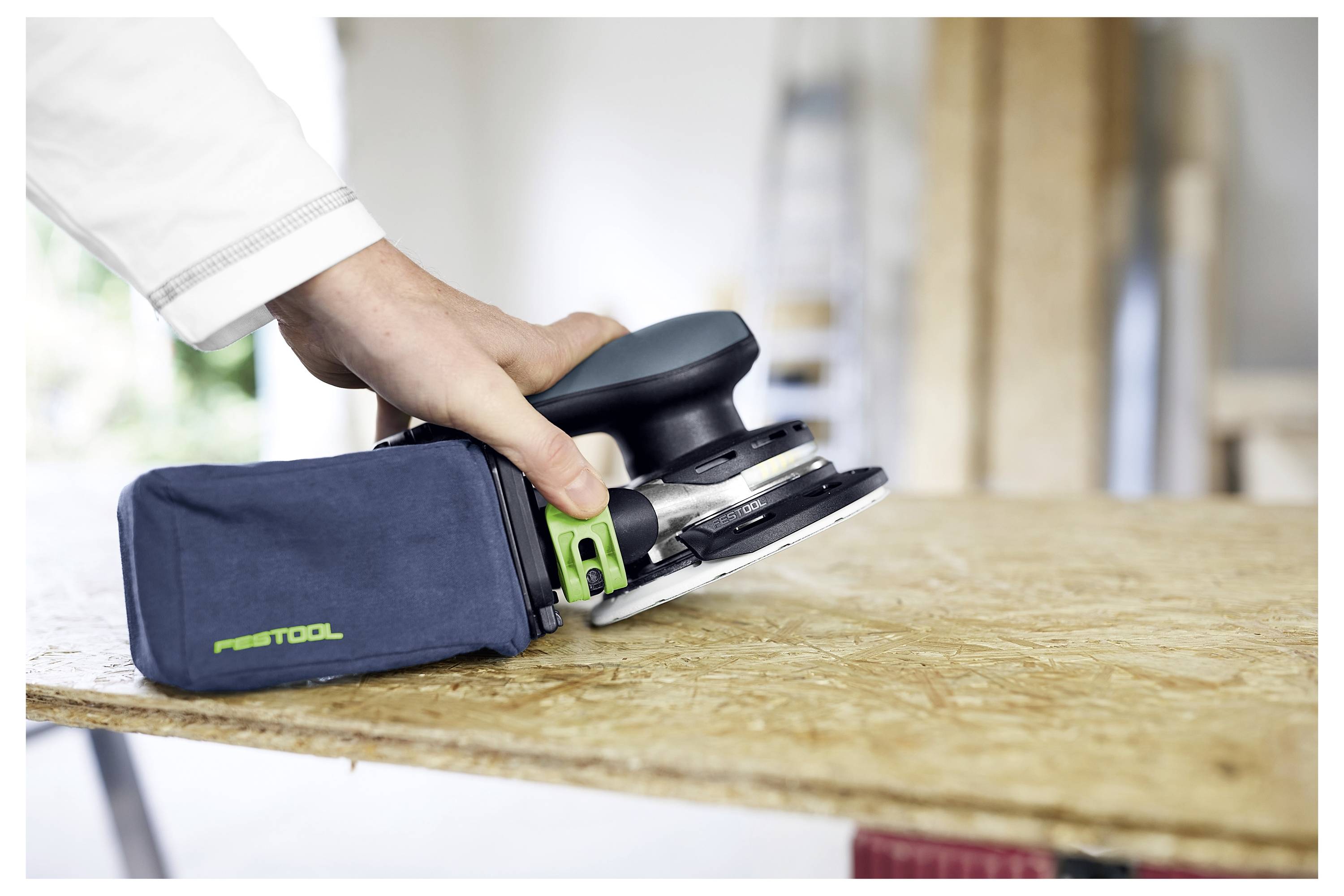 Eine Person in weißer Hose und grauem Hemd trägt einen Staubsauger und eine blaue Tasche in einem teilweise errichteten Gebäude.