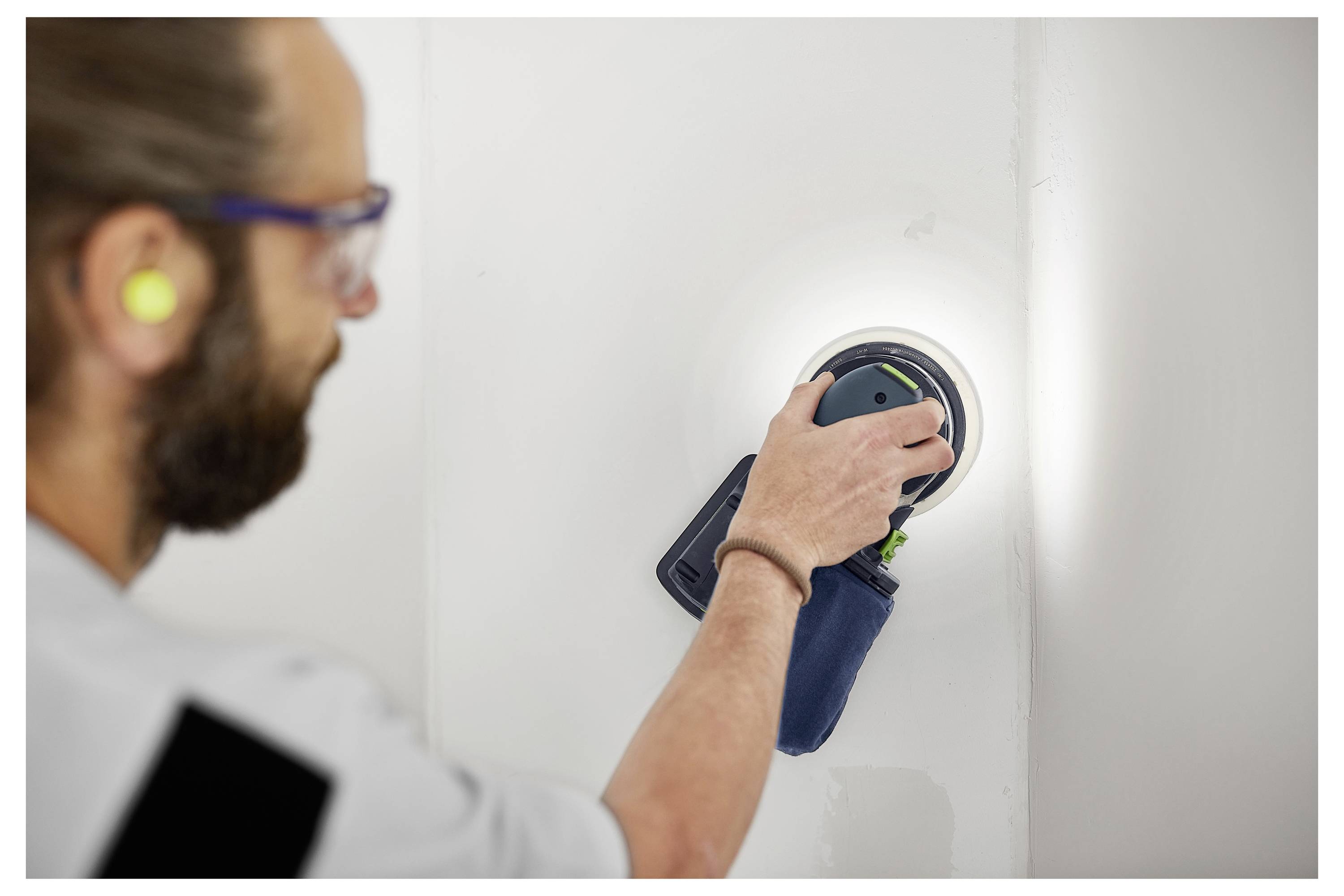 Eine Person trägt Ohrstöpsel und Schutzbrille und benutzt einen Elektroschleiffer an einer weißen Wand, wobei sie sich darauf konzentriert, die Oberfläche zu glätten.