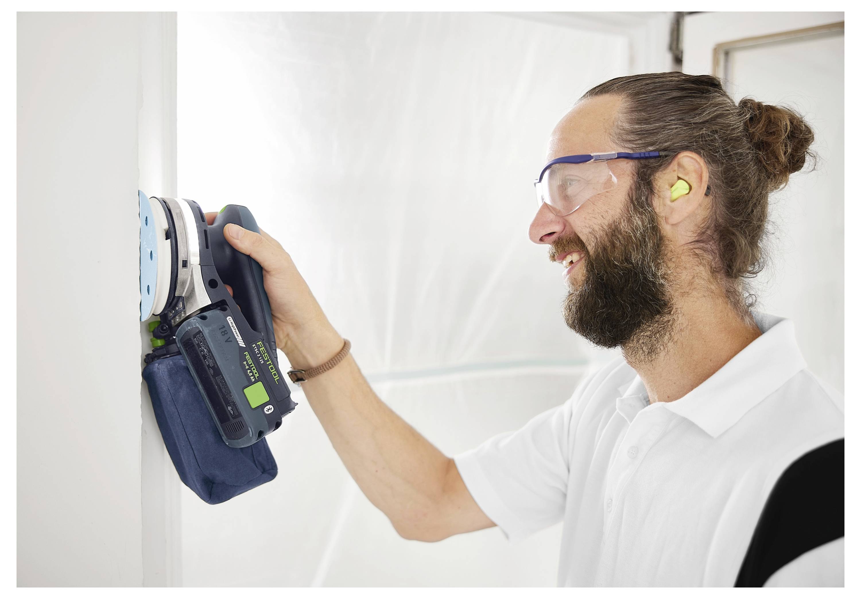 Eine Person, die eine Schutzbrille und einen Gehörschutz trägt, benutzt einen Elektroschleiffer an einer weißen Wand in einem Raum, der für Maler- oder Renovierungsarbeiten eingerichtet ist.