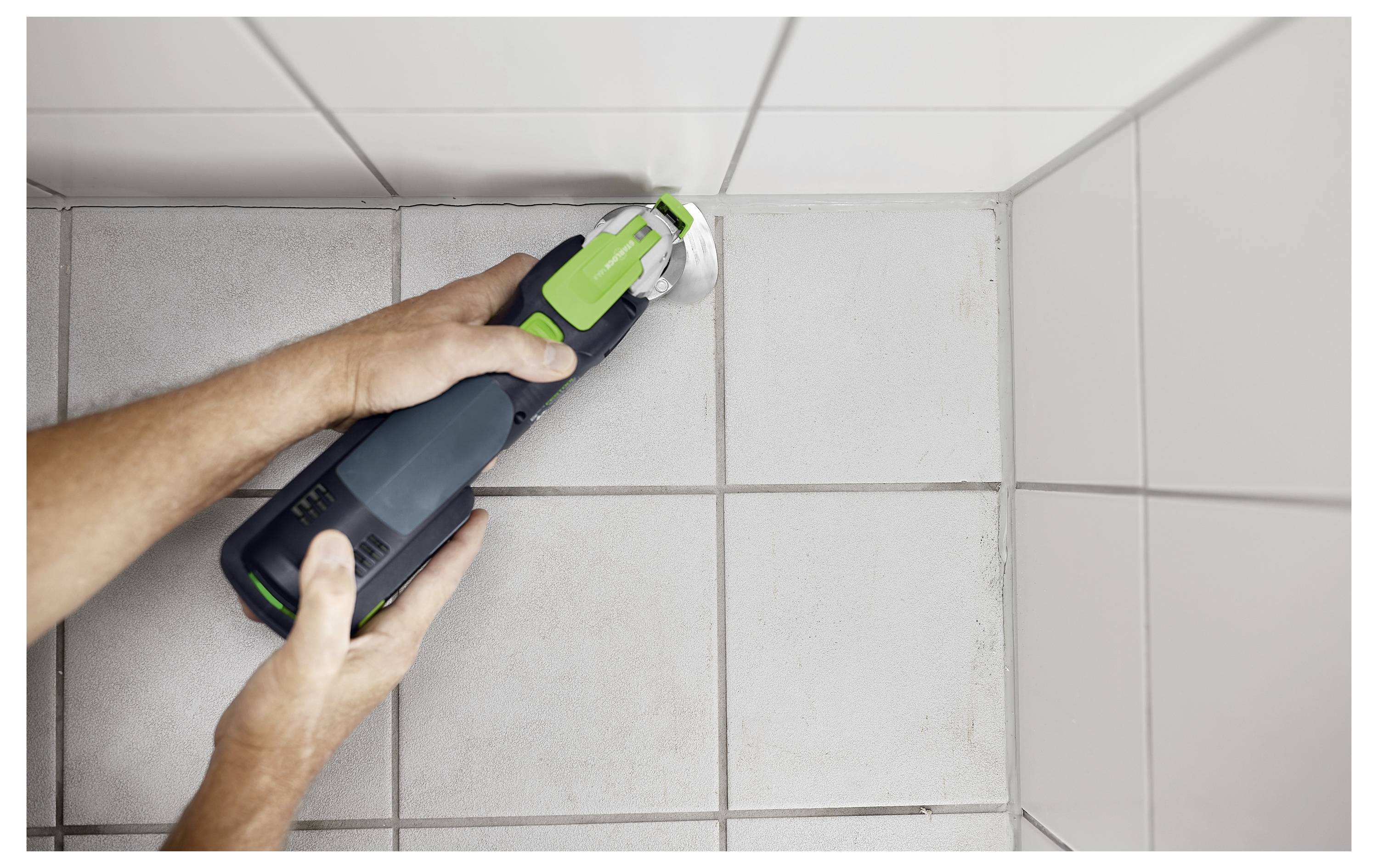 A person uses a handheld electric tool to cut ceramic tile grout in a tiled corner, focusing on precision and alignment.