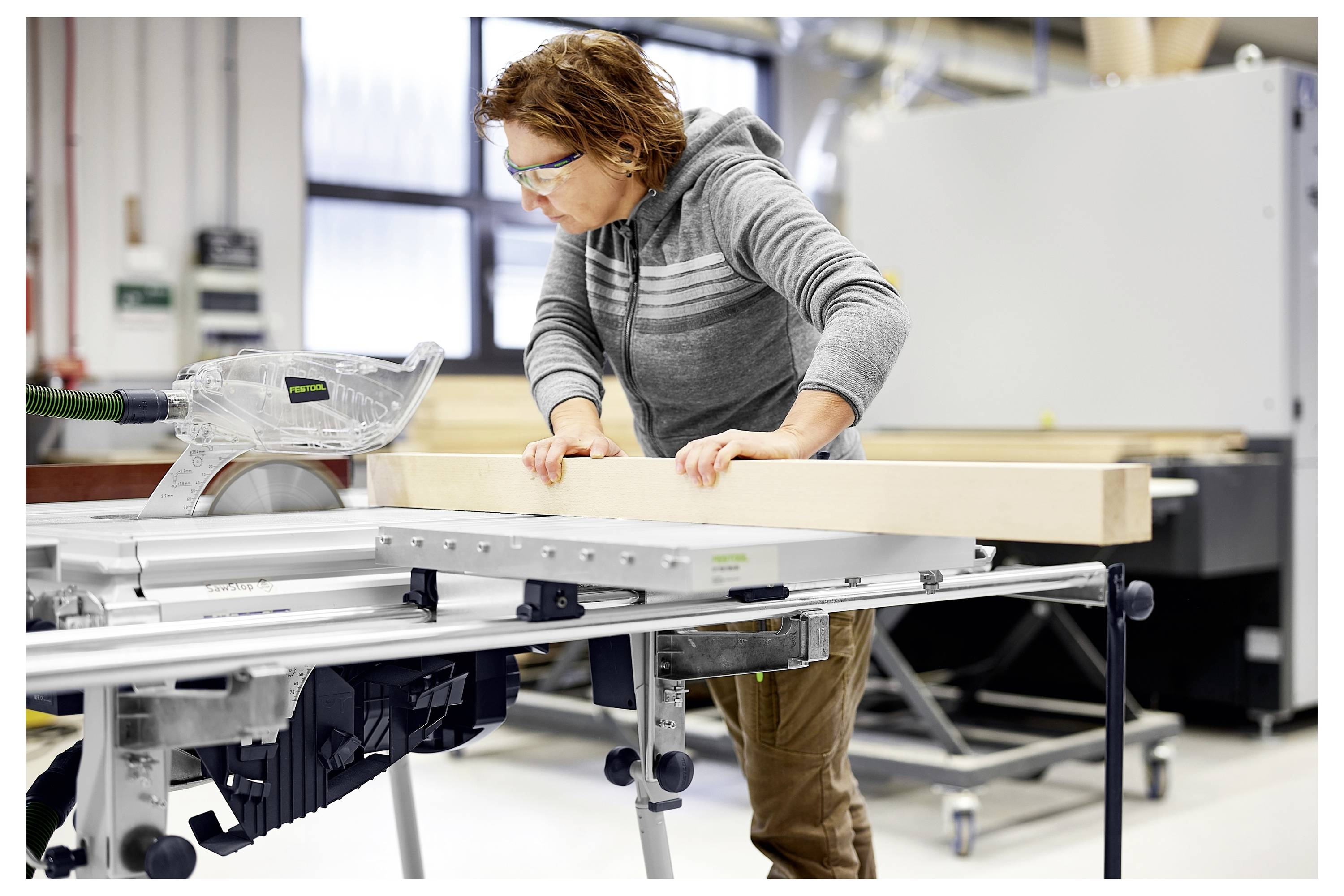 Eine Person mit Schutzbrille bedient eine Tischkreissäge und schneidet ein großes Holzstück in einer Werkstatt.