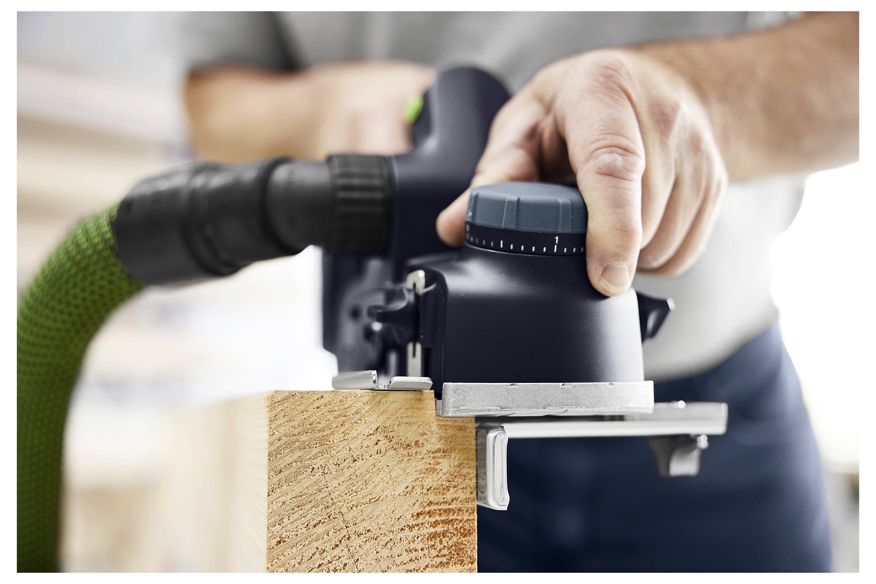 Person, die einen Elektroschleifers auf einem Holzbrett verwendet. Der Schleifers ist mit einem grünen Schlauch verbunden und glättet die Holzoberfläche.