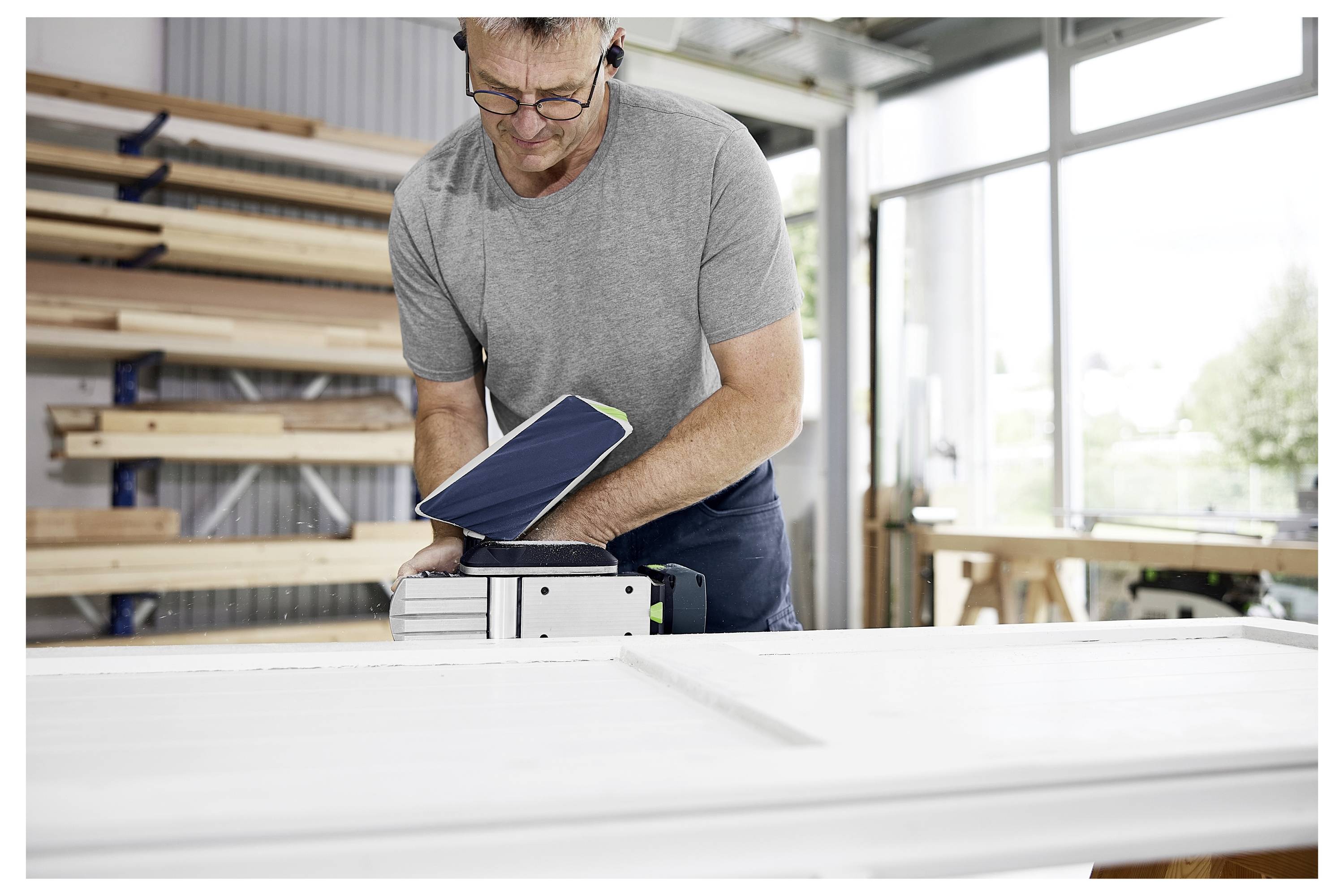 Eine Person in einer Werkstatt, die ein graues Hemd trägt, benutzt ein Elektrowerkzeug zum Schleifen einer weißen Holzoberfläche, mit Holzregalen im Hintergrund.