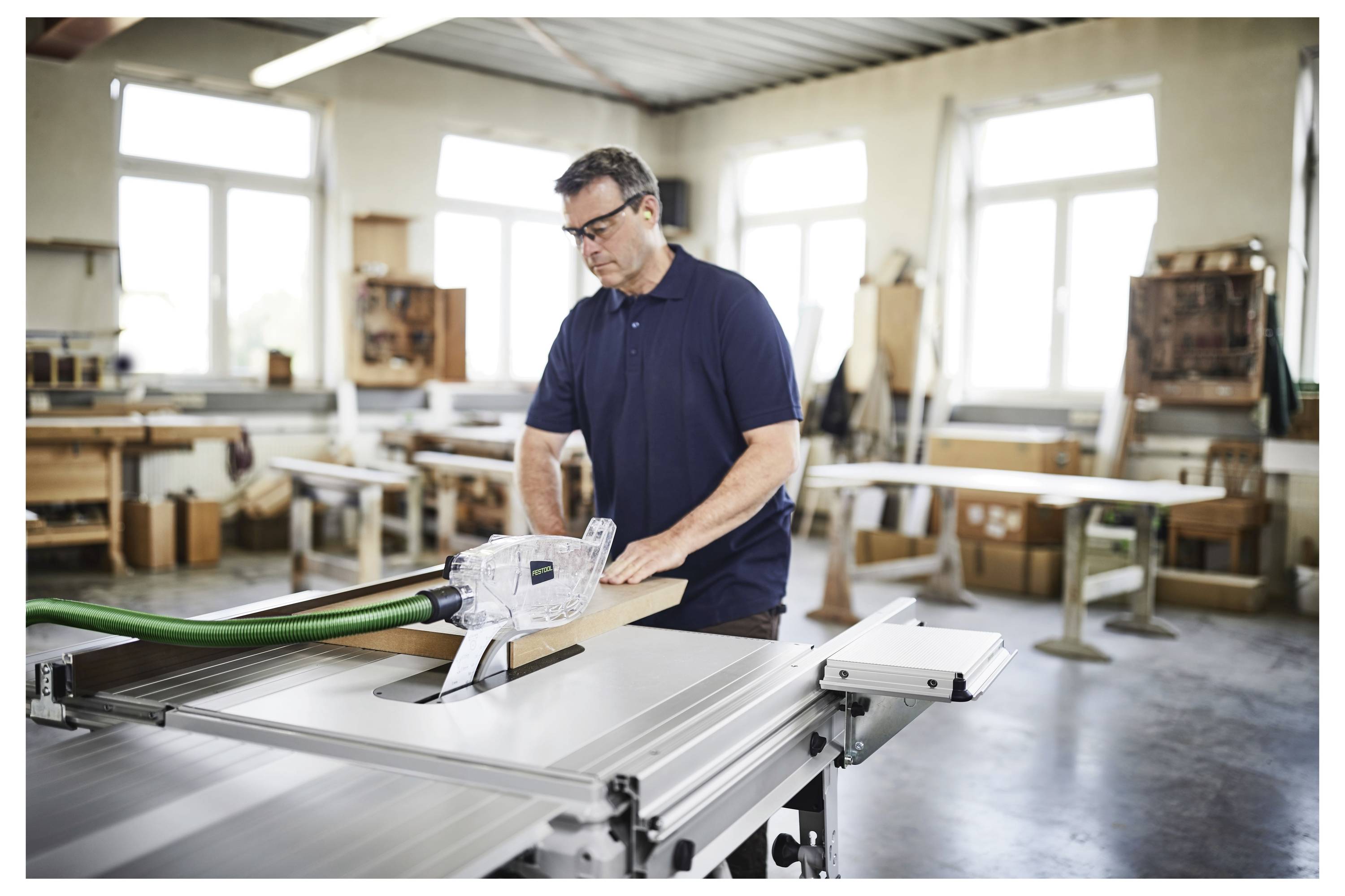Eine Person benutzt ein Elektrowerkzeug auf einem Holzbrett in einer Werkstatt. Sie trägt eine Schutzbrille und konzentriert sich auf ihre Arbeit in einer hellen, industriellen Umgebung.