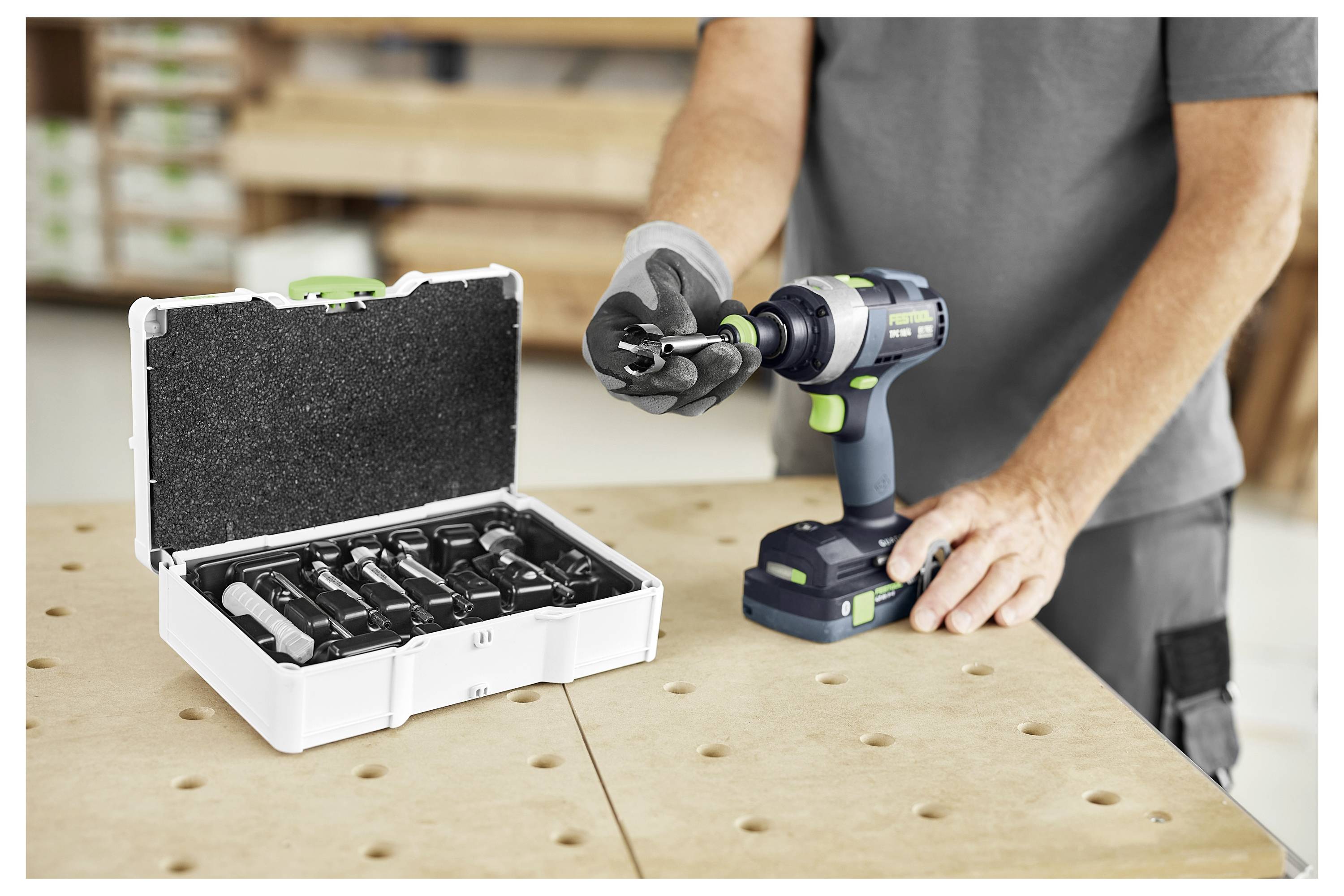 Eine Person, die Handschuhe trägt, benutzt einen kabellosen Bohrer mit einem offenen Bitinstallationssatz in der Nähe auf einer Werkbank in einer Werkstattumgebung.