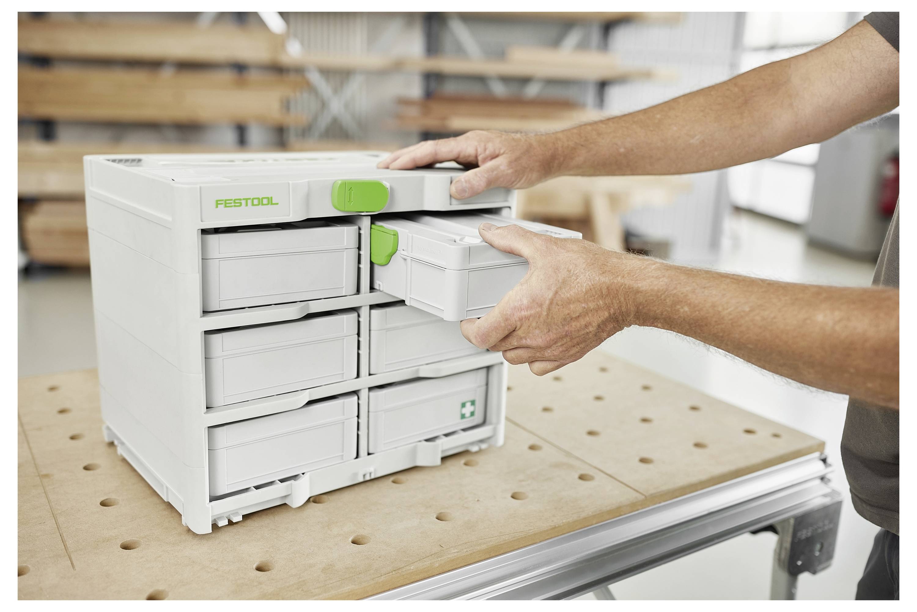 Ein Mann installiert Schränke in einem Raum. Der Fokus liegt auf einem Festool-Elektrowerkzeug, das auf einer Werkbank im Vordergrund platziert ist, mit einem angeschlossenen Schlauch.