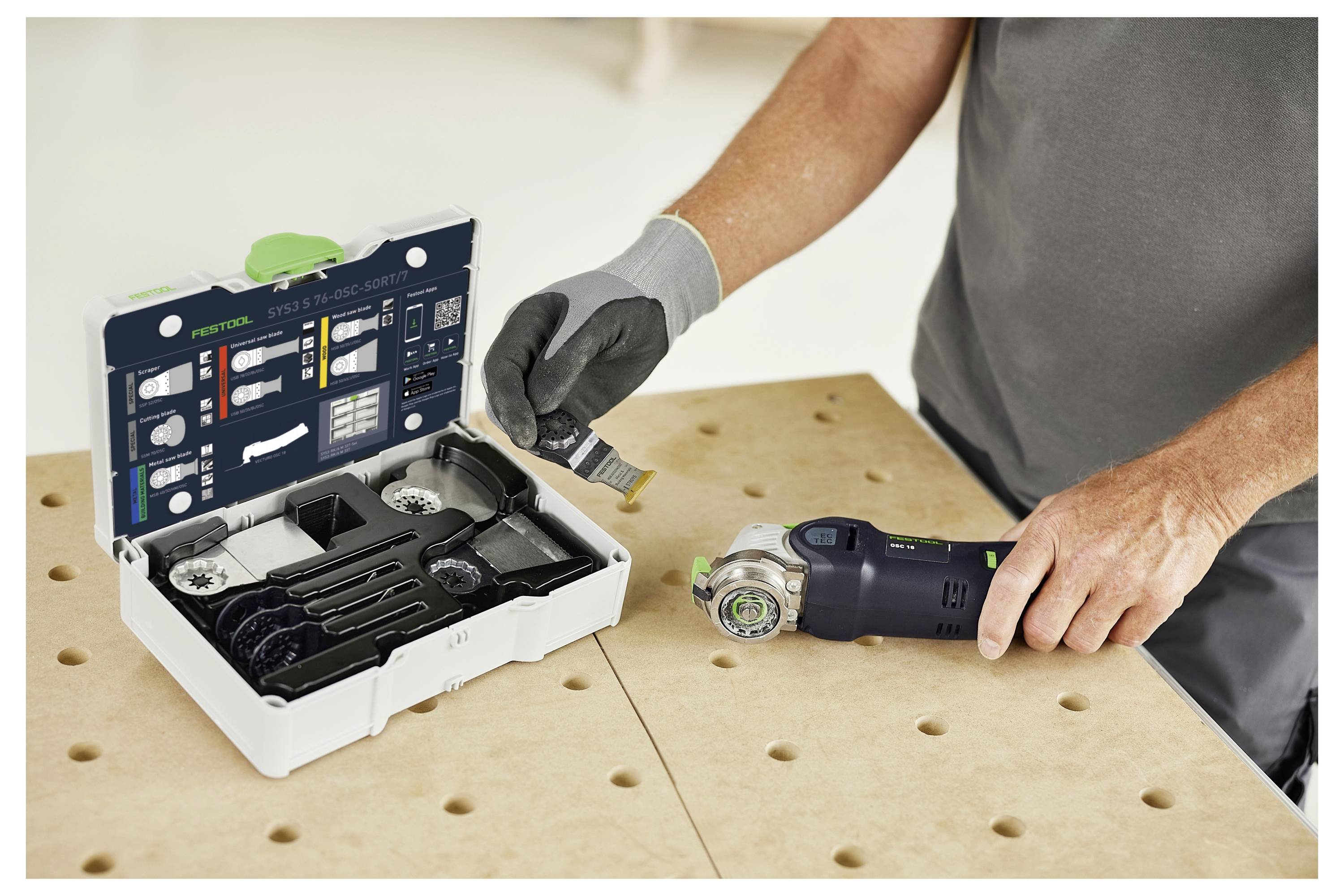 Eine Person nutzt ein kompaktes Elektrowerkzeug auf einer Sperrholzwerkbank neben einem offenen Koffer mit verschiedenen Werkzeugzubehörteilen.