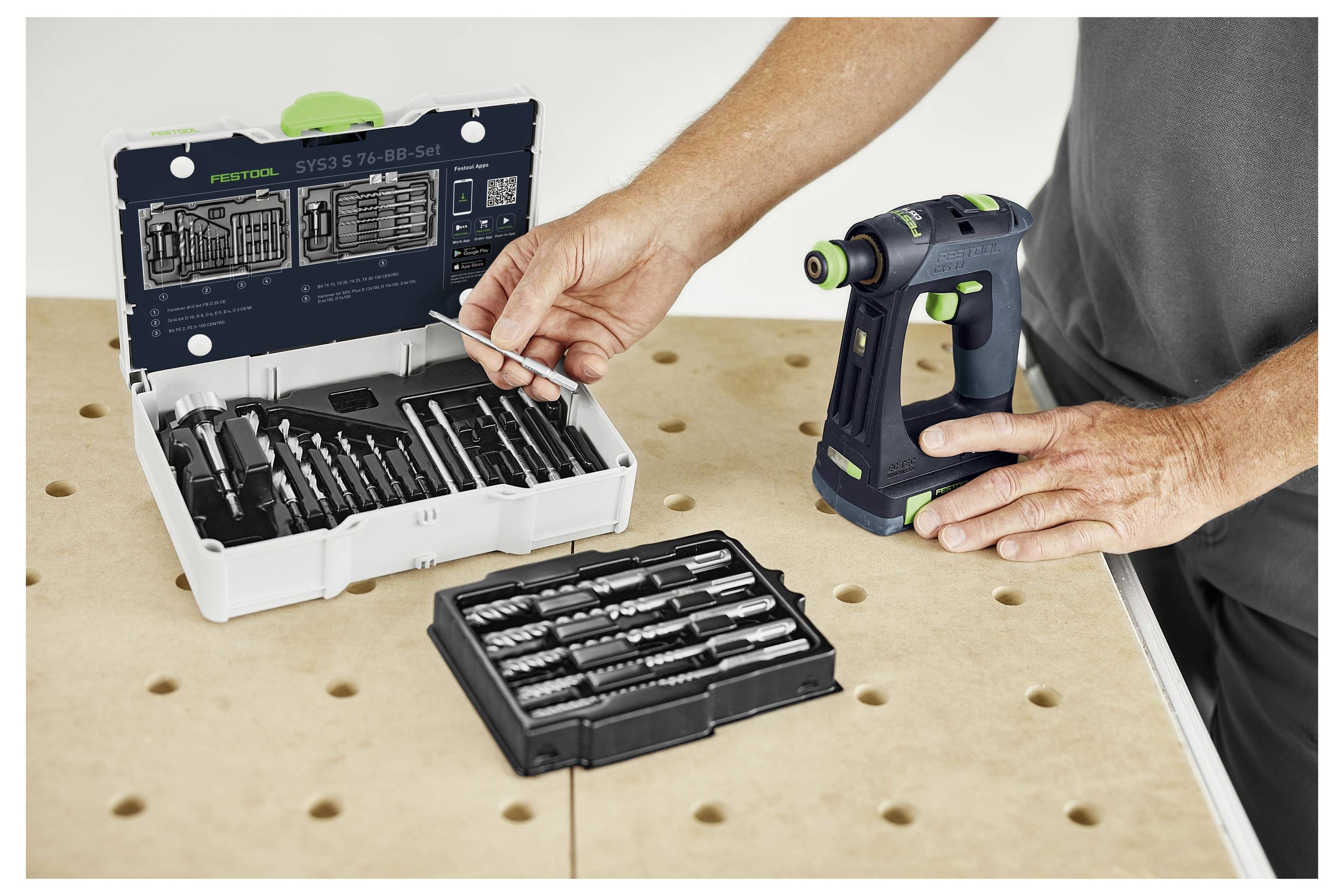 Eine Person wählt einen Bohrer aus einem Satz verschiedener Bohrer aus, die auf einer Werkbank liegen, neben denen sich ein Bohrwerkzeug befindet.