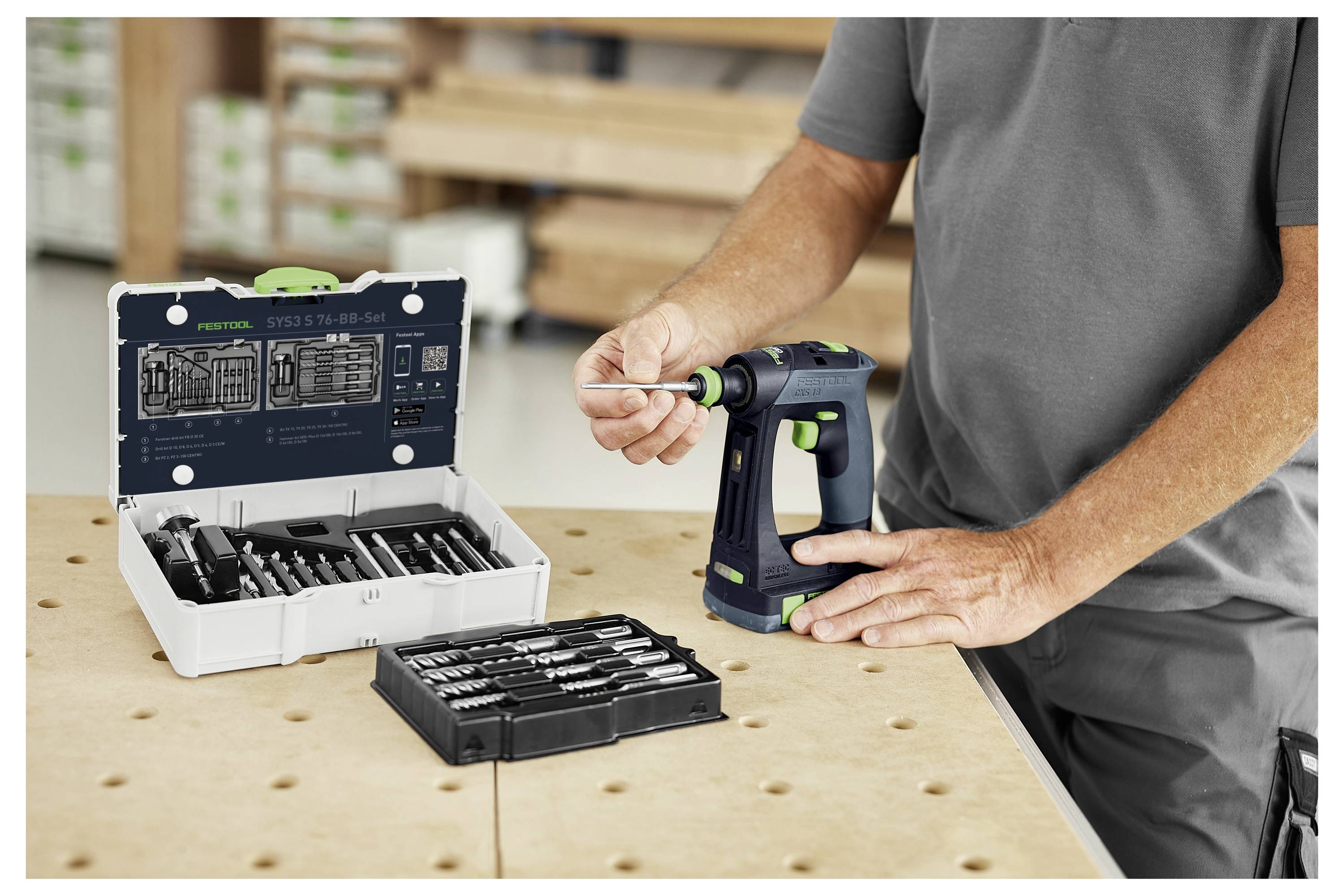 Eine Person bedient einen tragbaren Elektroschrauber neben einer offenen Werkzeugkiste mit verschiedenen Bohrern auf einer Werkbank in einer Werkstatt.