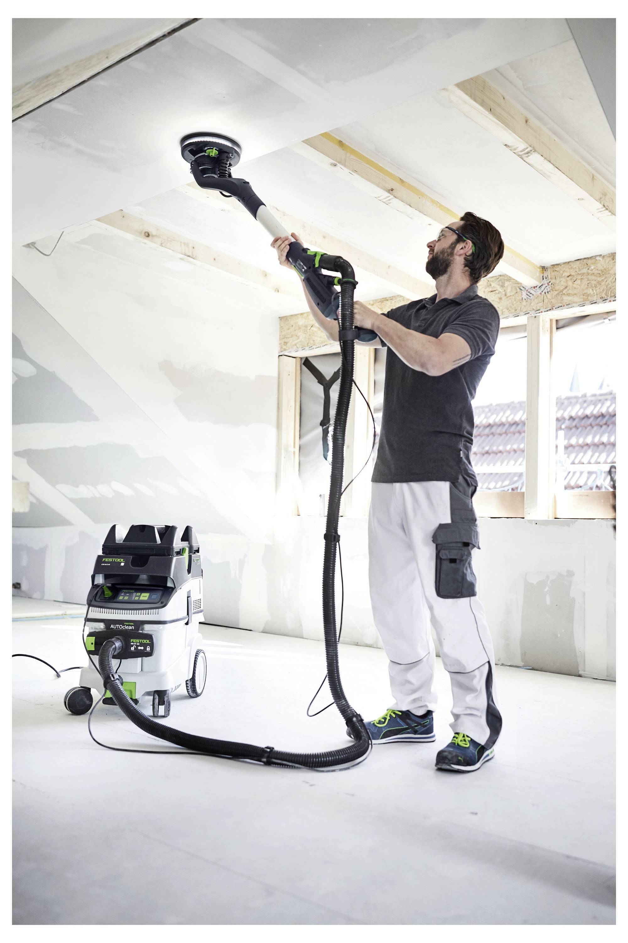 Eine Person benutzt einen Elektroschleiffer, um eine Decke in einem Baustelle zu glätten, mit einem Staubsauger, der an den Schleiffer angeschlossen ist, um Staub zu sammeln.