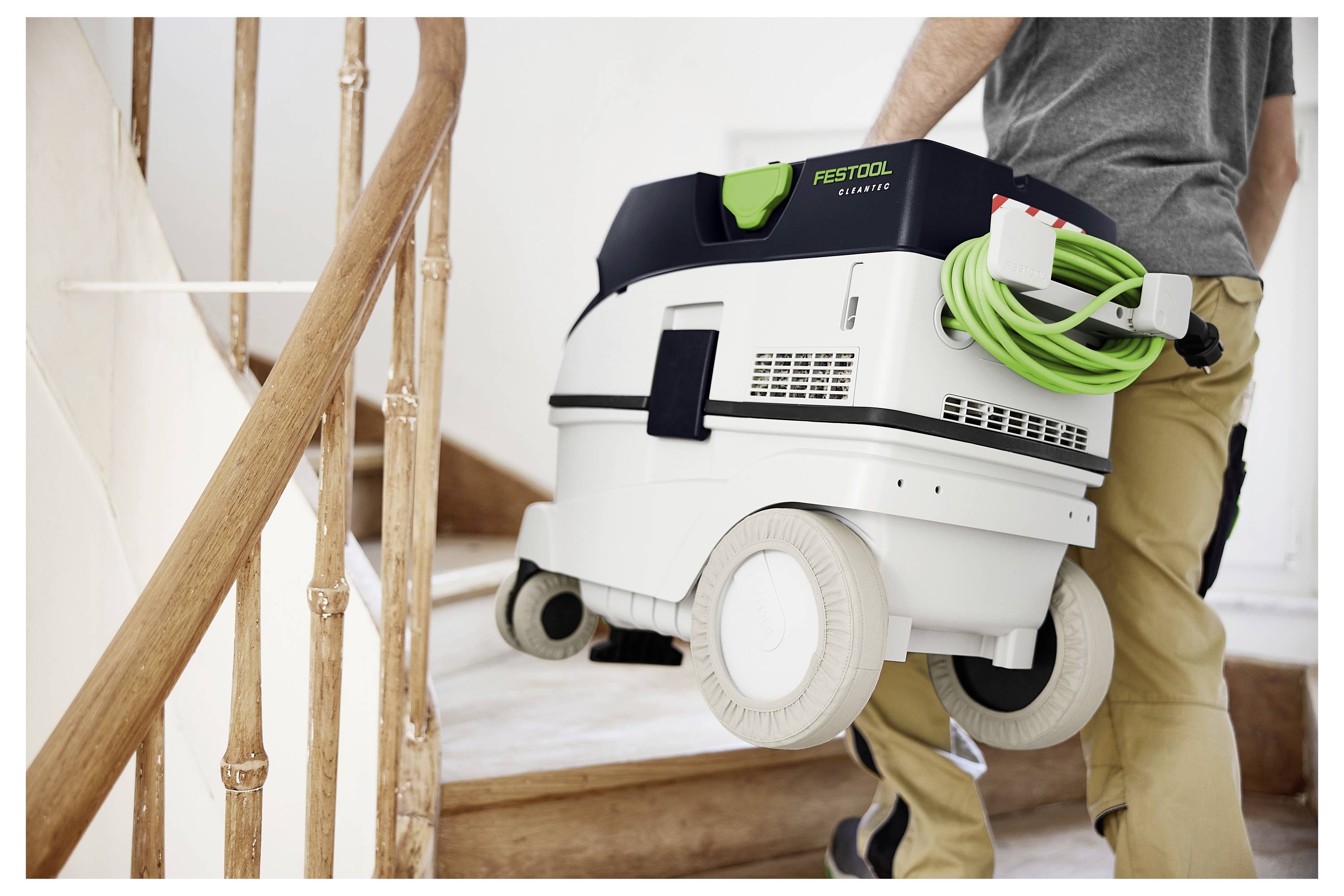 Eine Person in einem schwarzen Hoodie, lächelnd, steht in einer Holzwerkstatt mit Stapeln von Bauholz im Hintergrund.