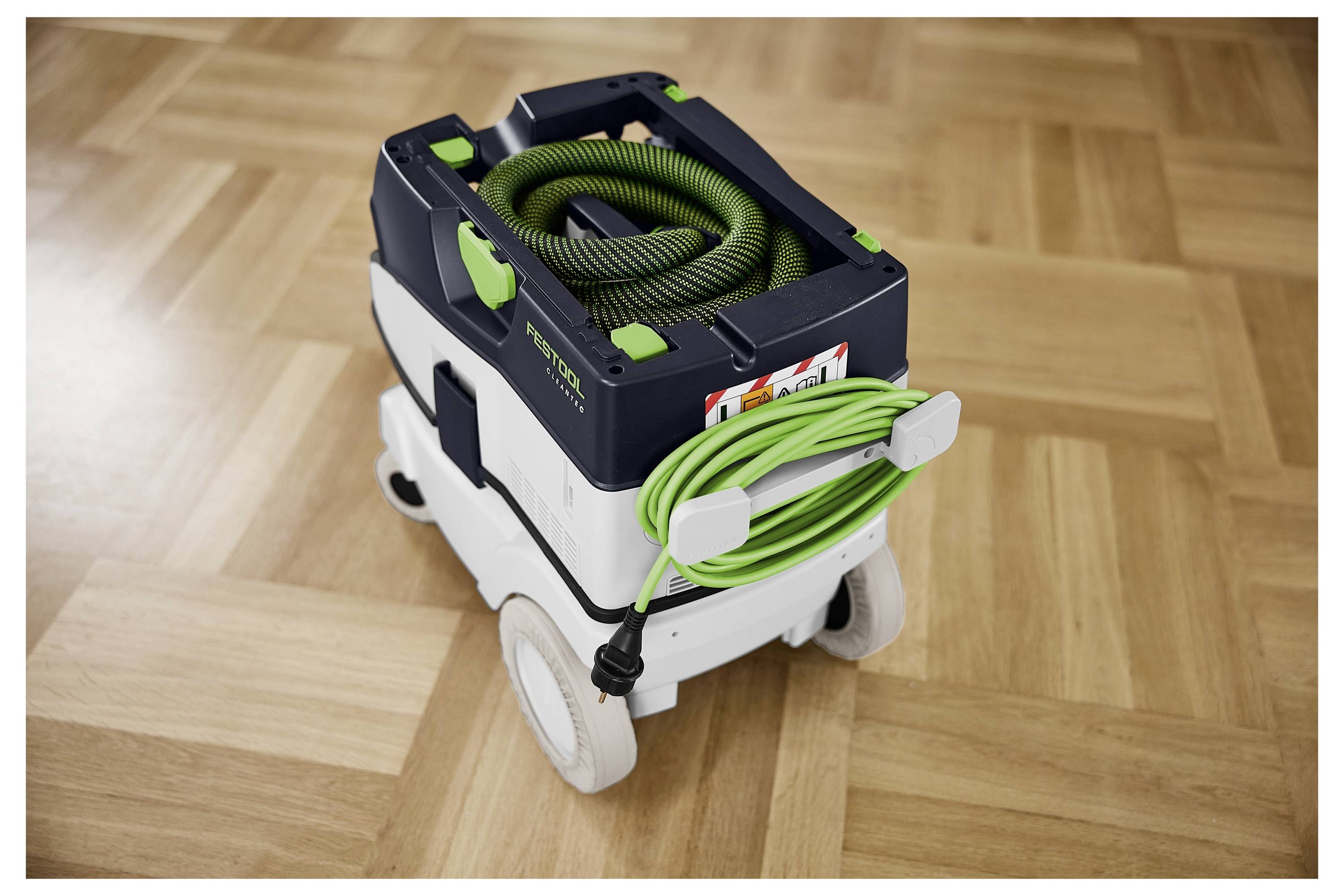 Ein tragbarer Staubsauger mit einem grünen Schlauch, der oben und an den Seiten aufgerollt ist, auf einem Holzboden, der sein kompaktes Design zur Schau stellt.