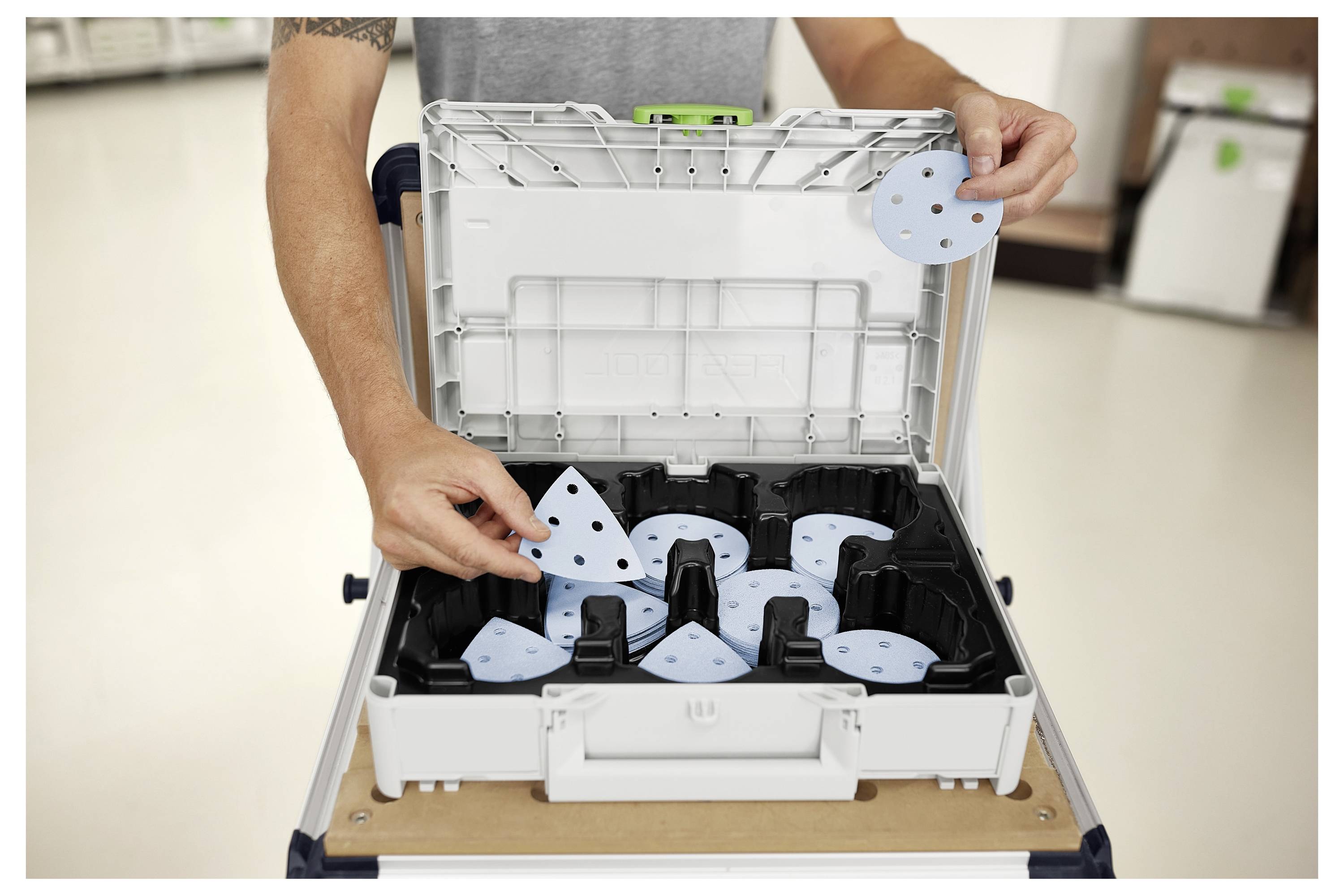 Ein Mann mit Schutzbrille benutzt eine Elektrosäge, um Holzbretter auf einer Baustelle zu schneiden, mit Gerüsten und Gebäuden im Hintergrund.