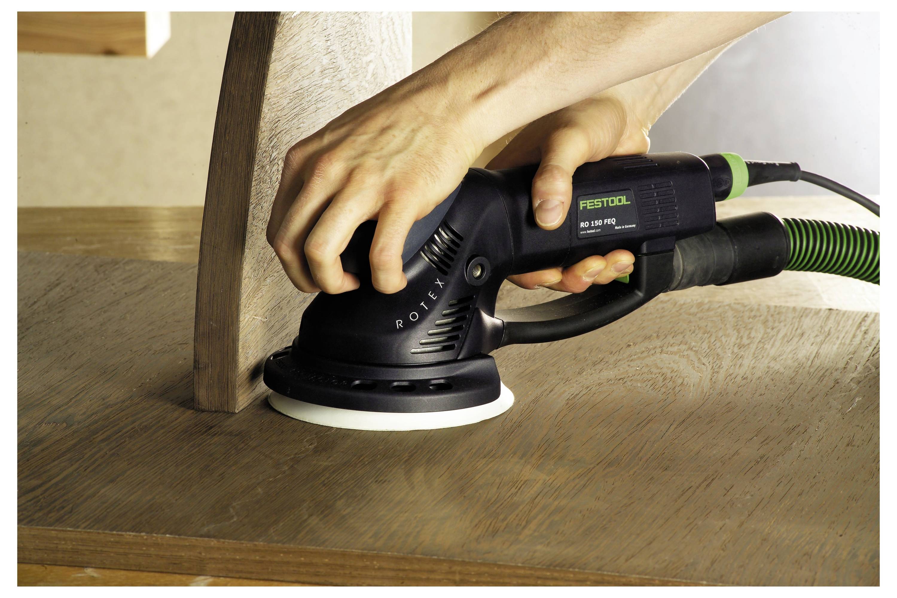 Eine Person benutzt einen elektrischen Schleiffer auf einer Holzoberfläche und glättet die Kanten für eine gleichmäßige Oberflächenbeschaffenheit. Der Schleiffer ist an einem grünen Schlauch befestigt.