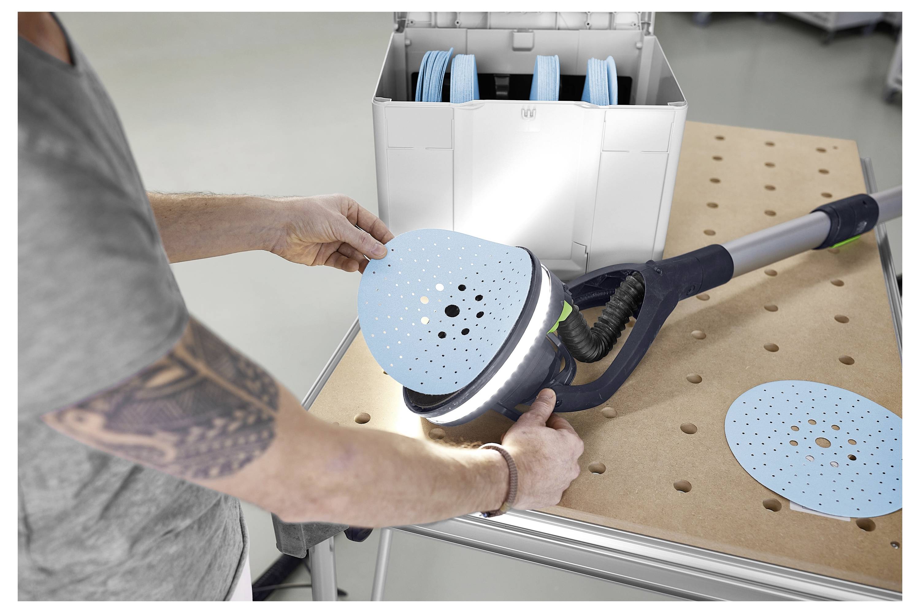 Eine Person schleift Trockenbau in einem Renovierungszimmer mit einem an einen Staubsauger angeschlossenen Elektroschleiffer ab, umgeben von unfertigen Wänden.