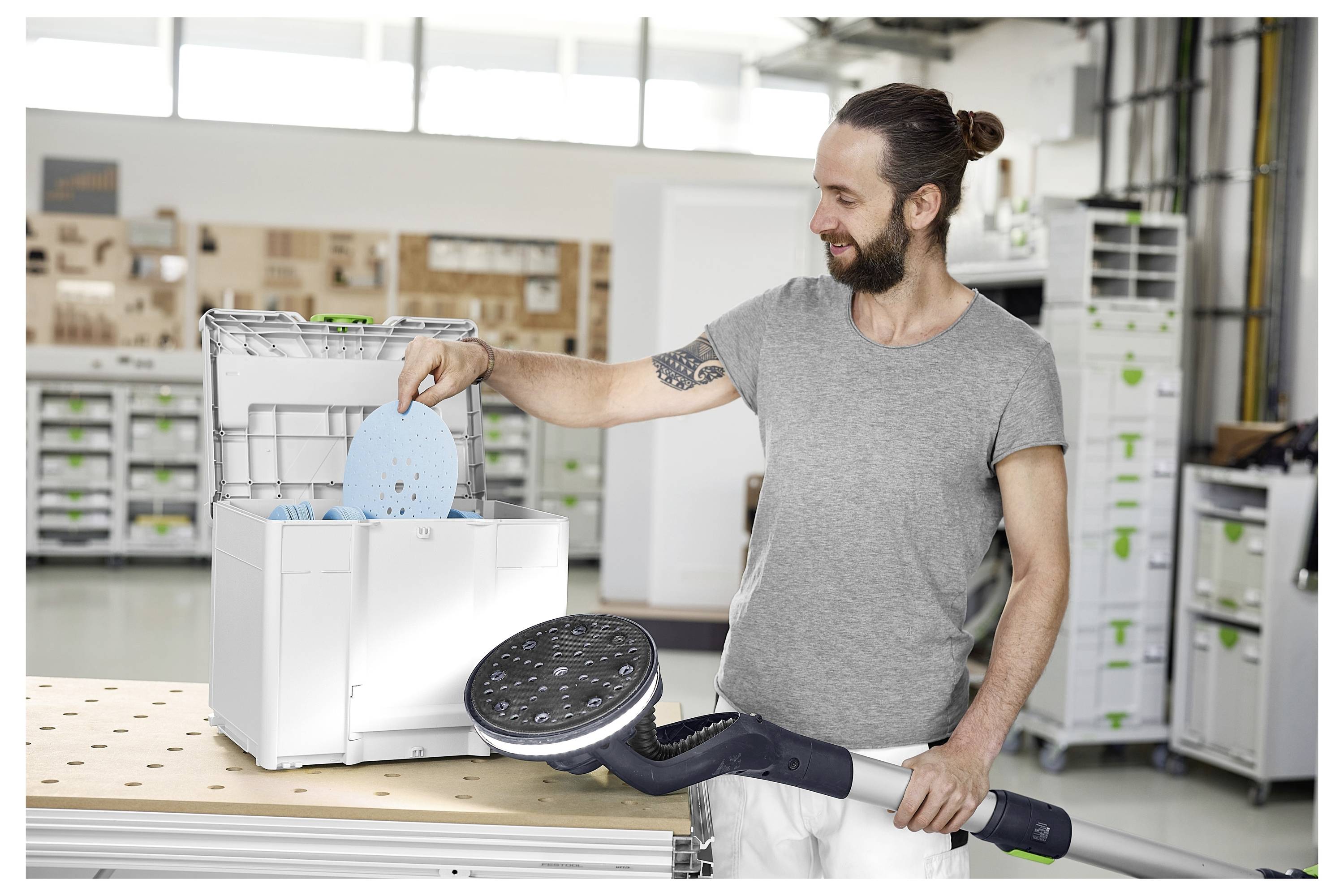 Ein Mann in einer Werkstatt, der ein großes Elektrowerkzeug hält und runde Schleifmittelscheiben in eine Aufbewahrungsbox lädt. Im Hintergrund befinden sich Regale und Ausrüstung.