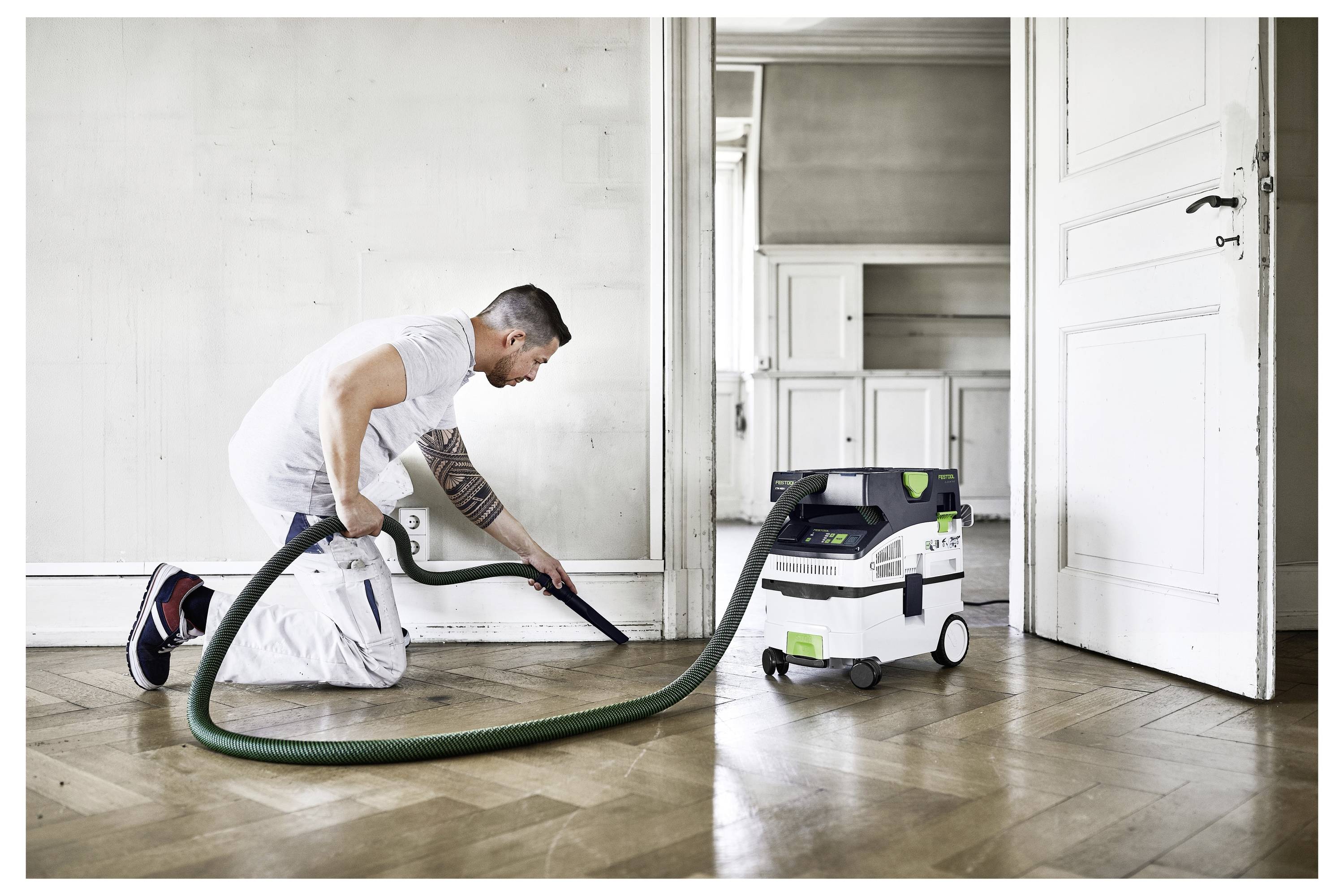 Eine Person benutzt einen Staubsauger auf einem Holzboden in einem Zimmer mit weißen Wänden und offenen Türen, was auf Reinigung oder Hauswartung hindeutet.