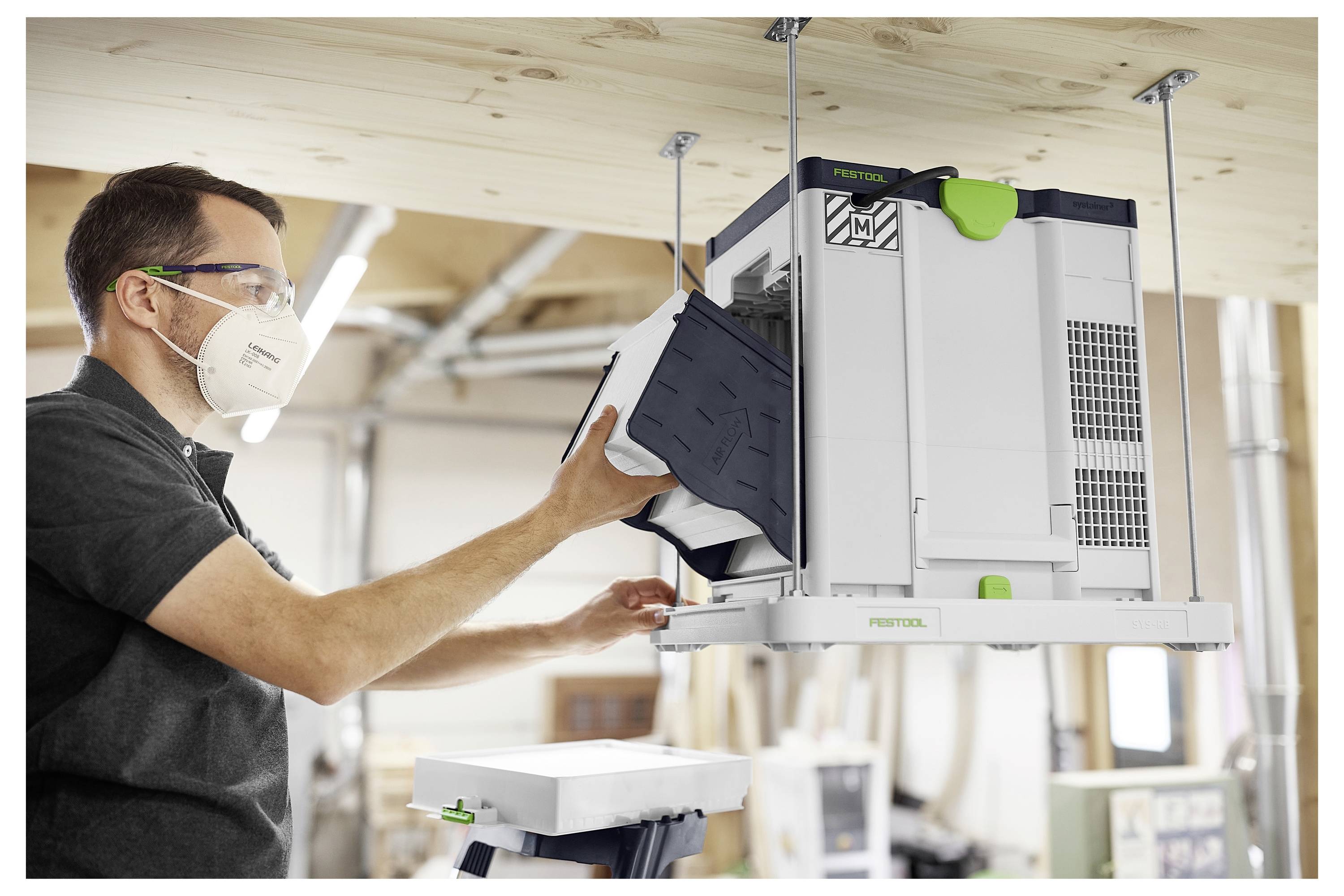 Eine Person installiert ein Lüftungssystem an einer Holzdecke in einer Werkstatt und trägt dabei eine Schutzbrille und eine Maske zu ihrer Sicherheit.