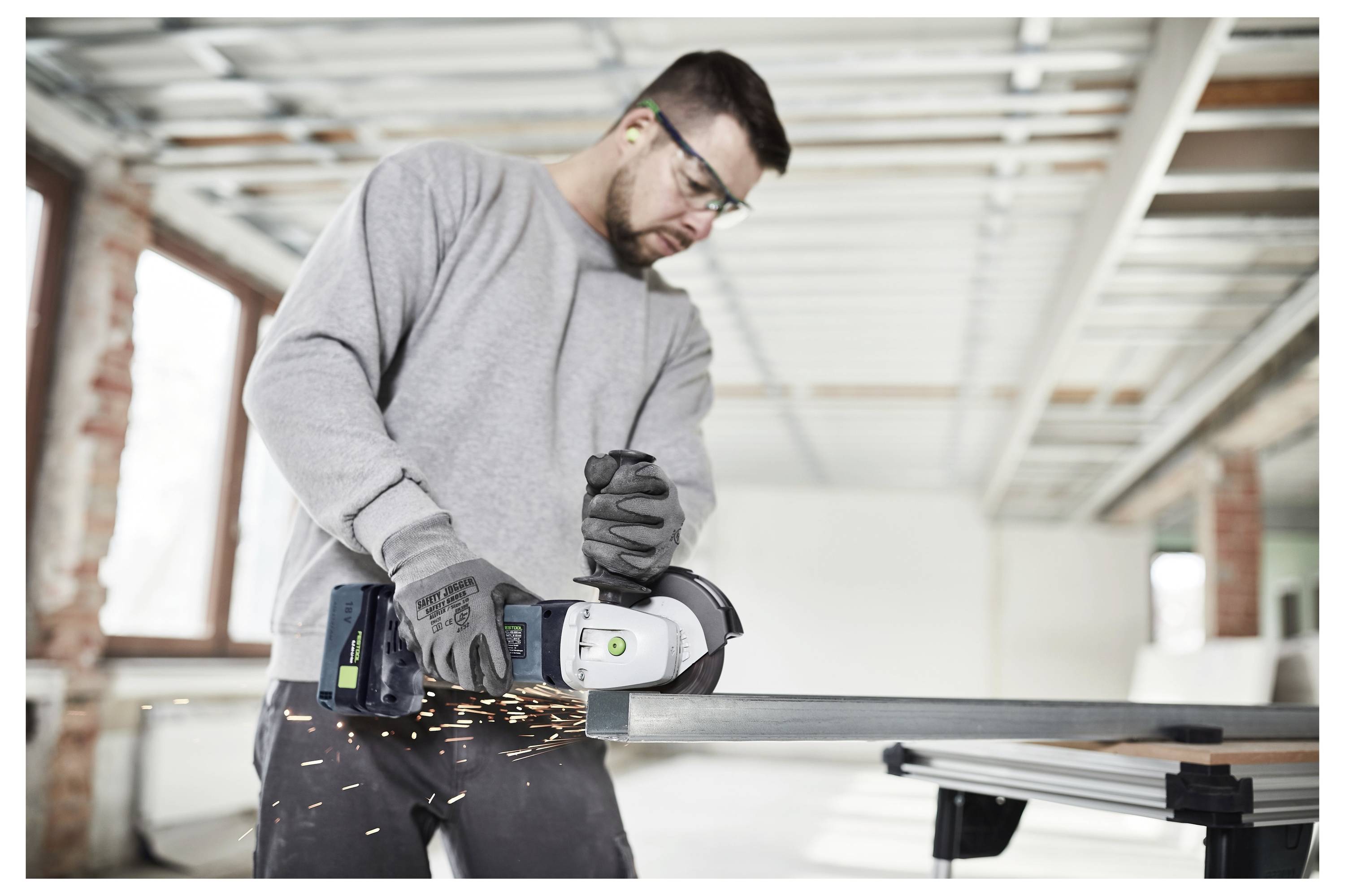Eine Person kniet auf einer Baustelle und benutzt ein Elektrowerkzeug, das mit einem Staubabsauger verbunden ist. Das Gerät steht im Mittelpunkt und unterstreicht die Staubkontrolle.