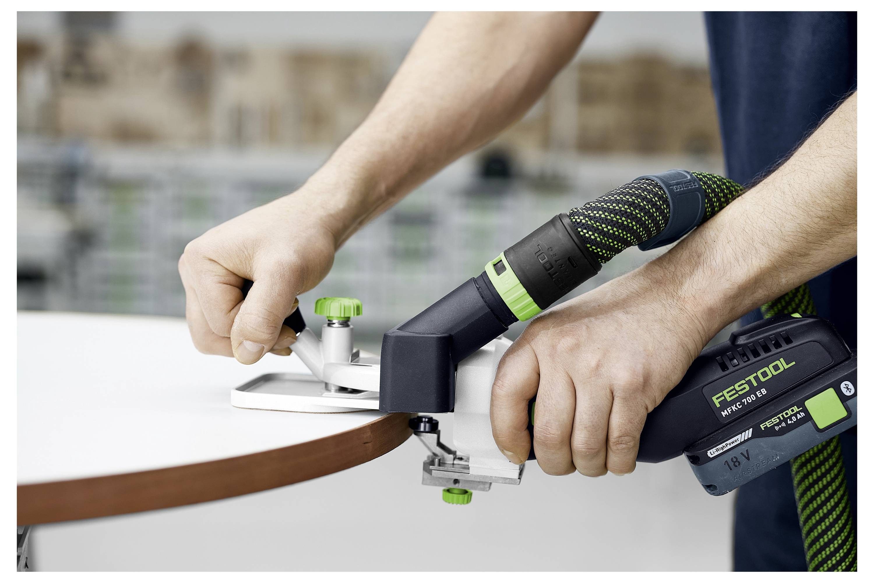 Eine Person verwendet ein kabelloses Kantenbandgerät, um eine Kantenleiste auf ein Holzbrett aufzubringen. Das Gerät ist grün und schwarz und trägt die Marke 