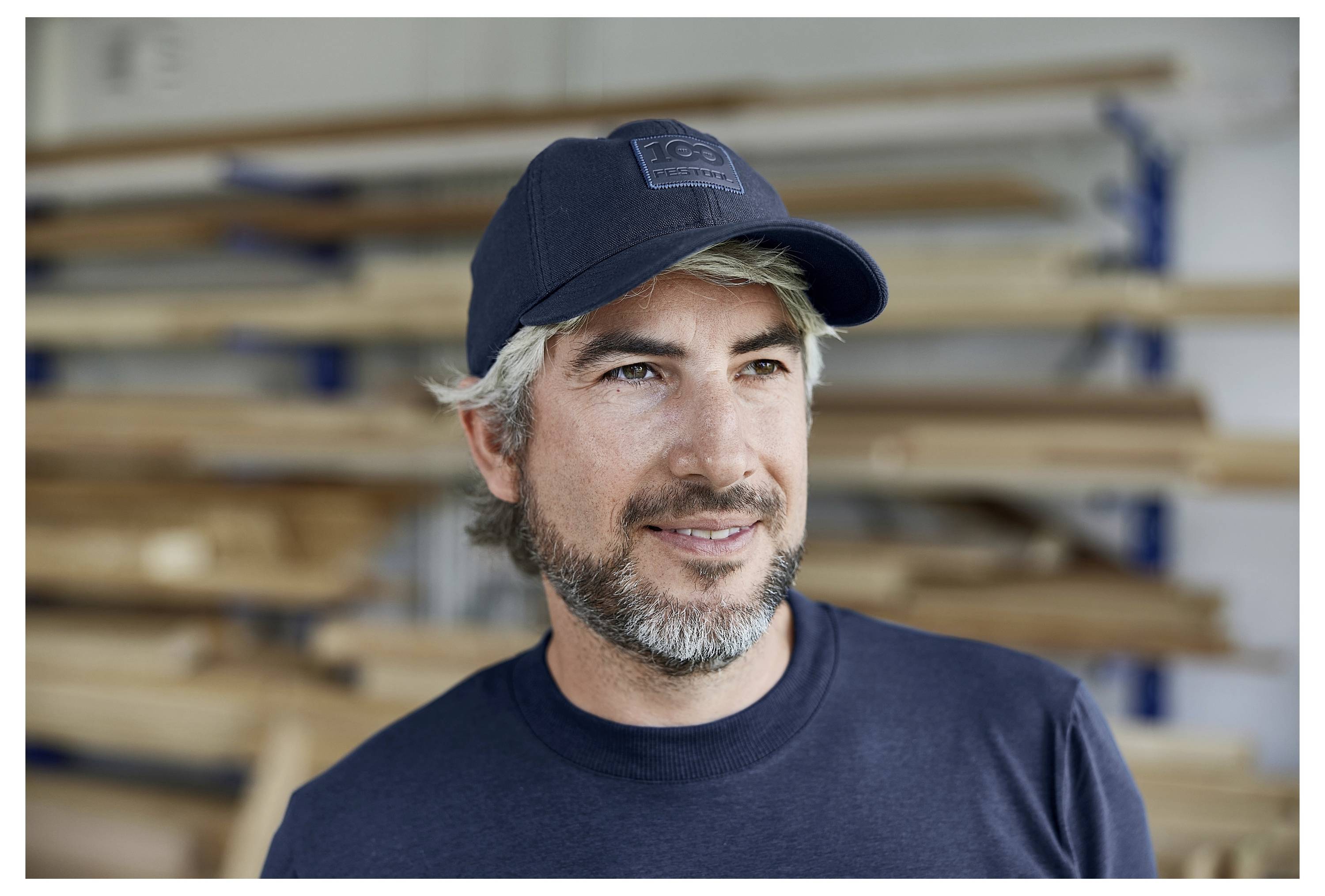 Ein Mann mit Bart und dunkeler Mütze steht in einer Werkstatt mit gestapelten Holzbrettern im Hintergrund.