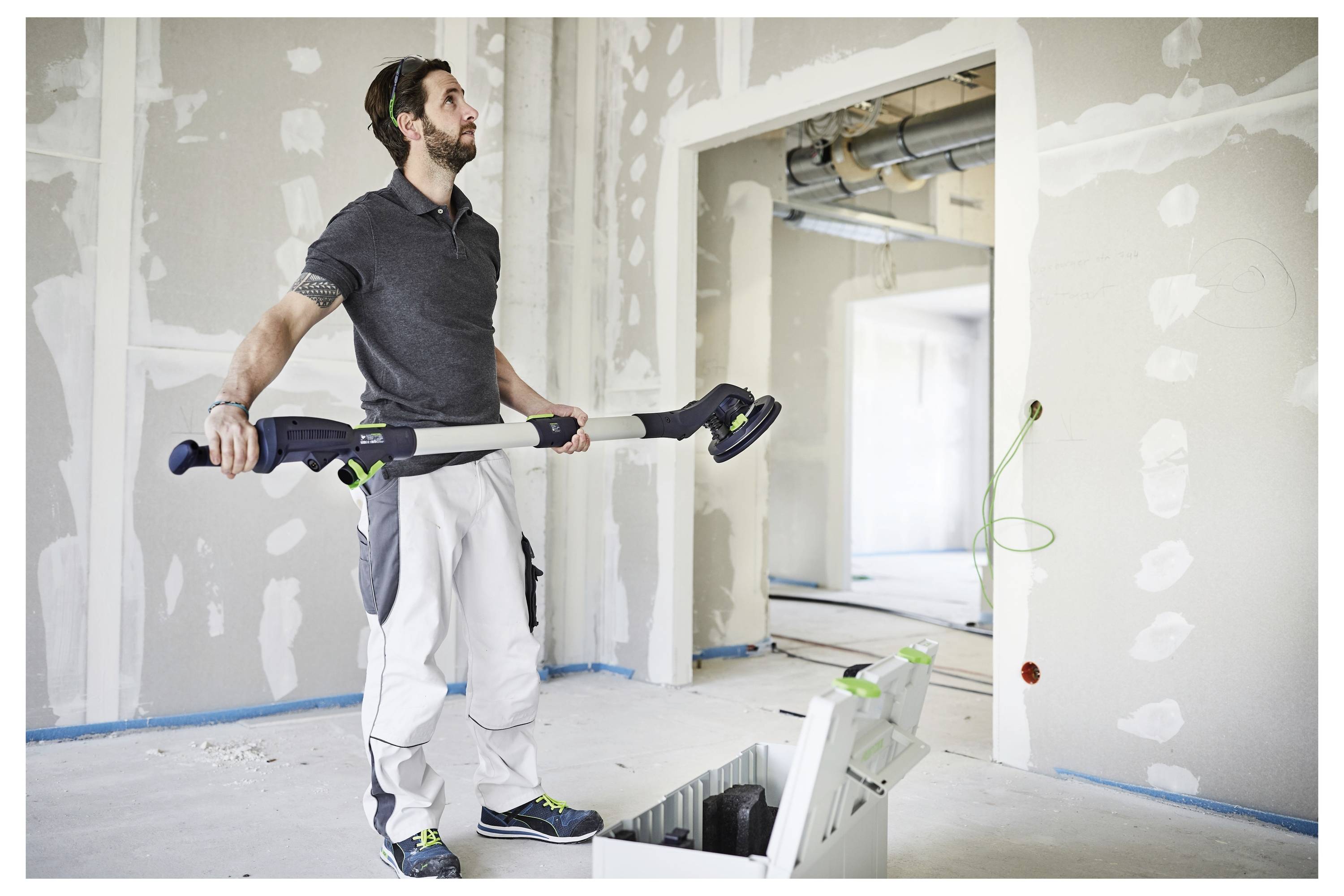 Eine Person in Schutzbrille verwendet einen elektrischen Schleiffer an einem Deckenbalken und glättet die Oberfläche. Sie trägt ein Sweatshirt und einen Gehörschutz.