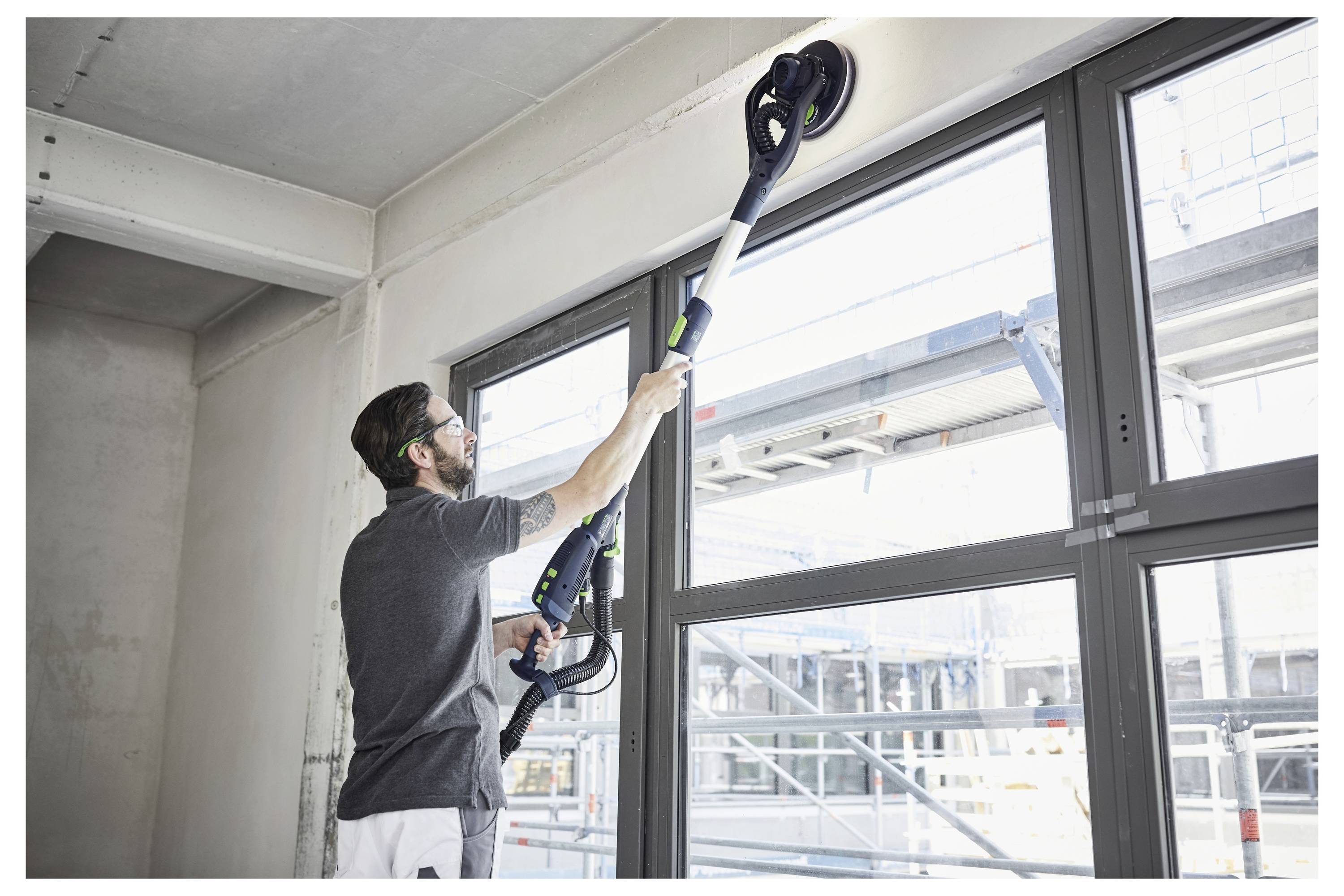Person, die ein Elektroschleifergerät verwendet, um eine hohe Wand in der Nähe großer Fenster in einem unfertigen Gebäude zu schleifen, was auf Bau- oder Renovierungsarbeiten hindeutet.
