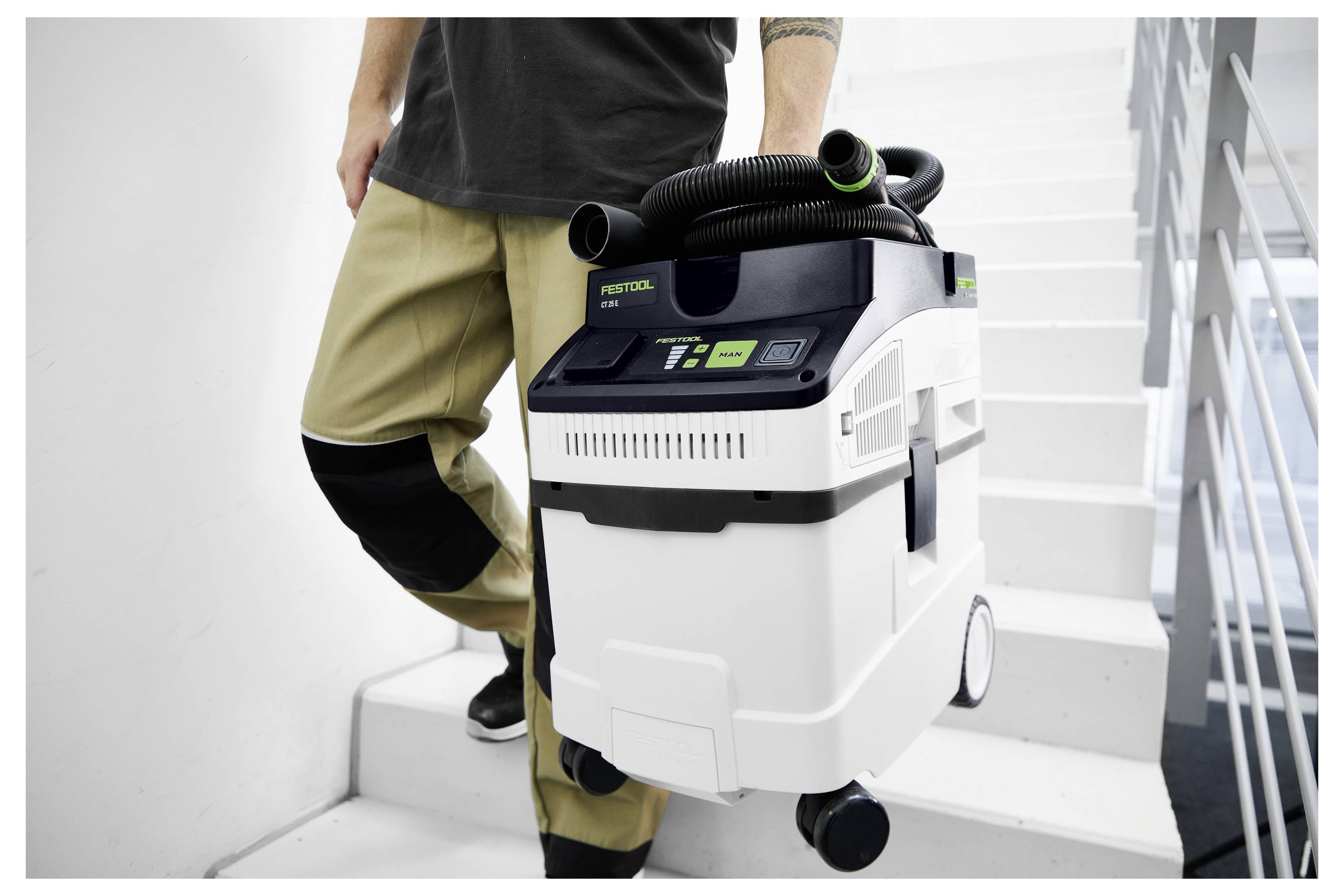 Eine Person trägt einen fahrbaren, kompakten Staubsauger eine Treppe hinauf, was die Tragbarkeit und Benutzerfreundlichkeit des Staubsaugers beim Reinigen von Treppen verdeutlicht.