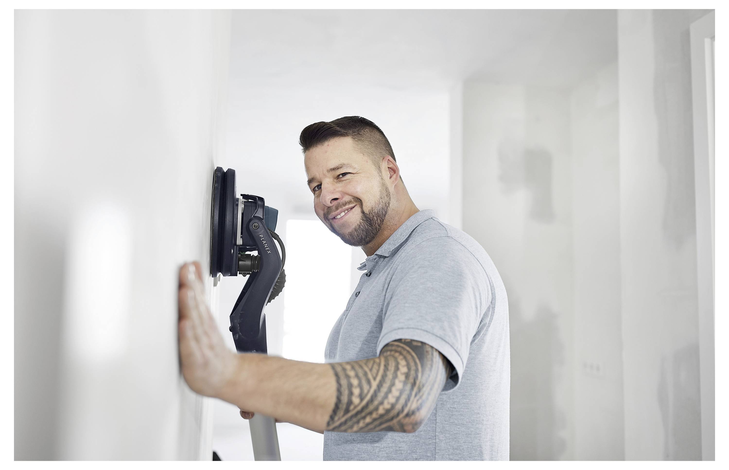 Mann, der einen Wandschlifer an einer weißen Wand verwendet, trägt ein hellgraues Poloshirt, lächelt, mit einem tätowierten Arm, in einem teilweise renovierten Zimmer.