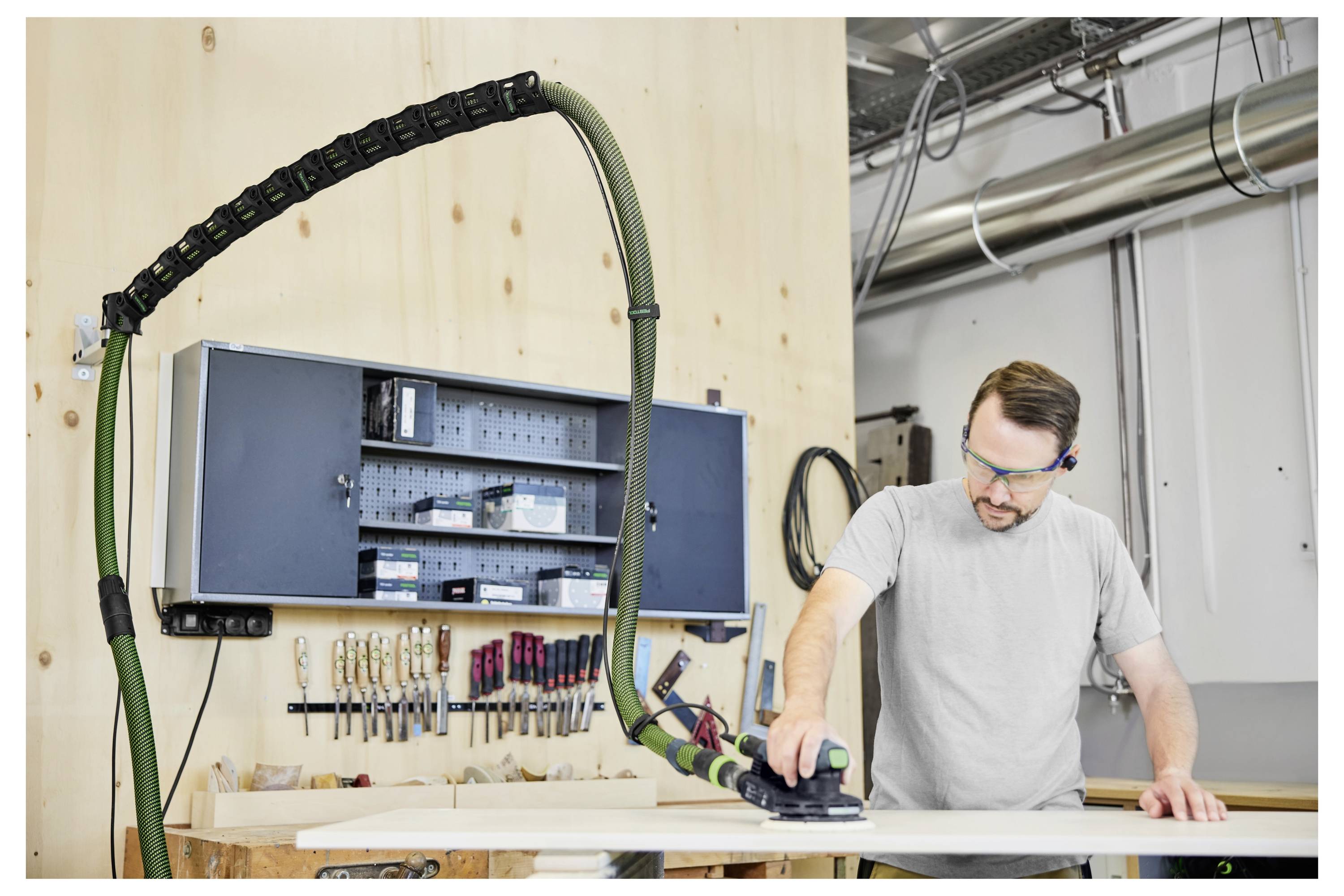 Eine Person, die ein Elektrowerkzeug verwendet, um ein Materialblatt zu schneiden, trägt eine Schutzbrille. Im Hintergrund sind Gerüste und ein Gebäude sichtbar.