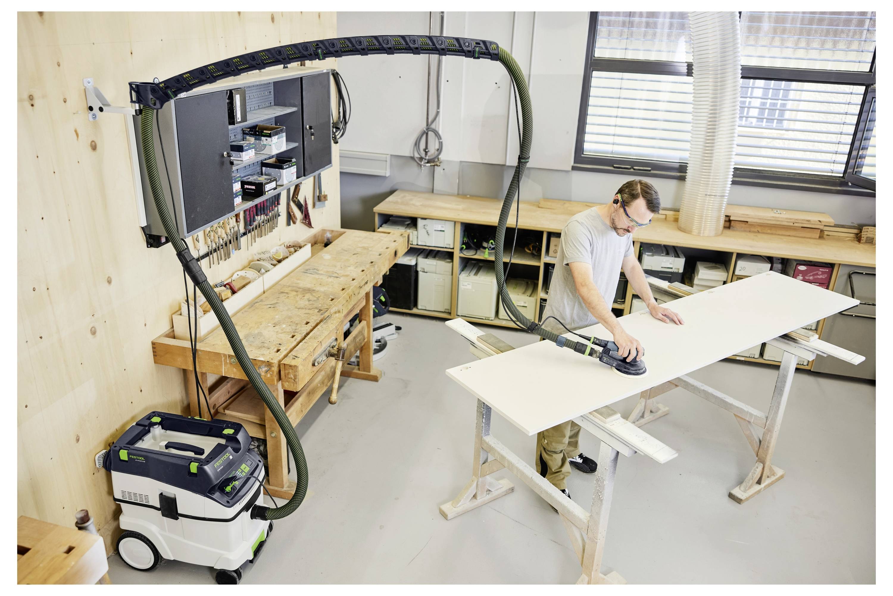 Ein Mann mit einer Mütze und einem Langarmhemd steht in einer Holzwerkstatt und blickt auf Regale, die mit gestapelten Holzbrettern gefüllt sind.