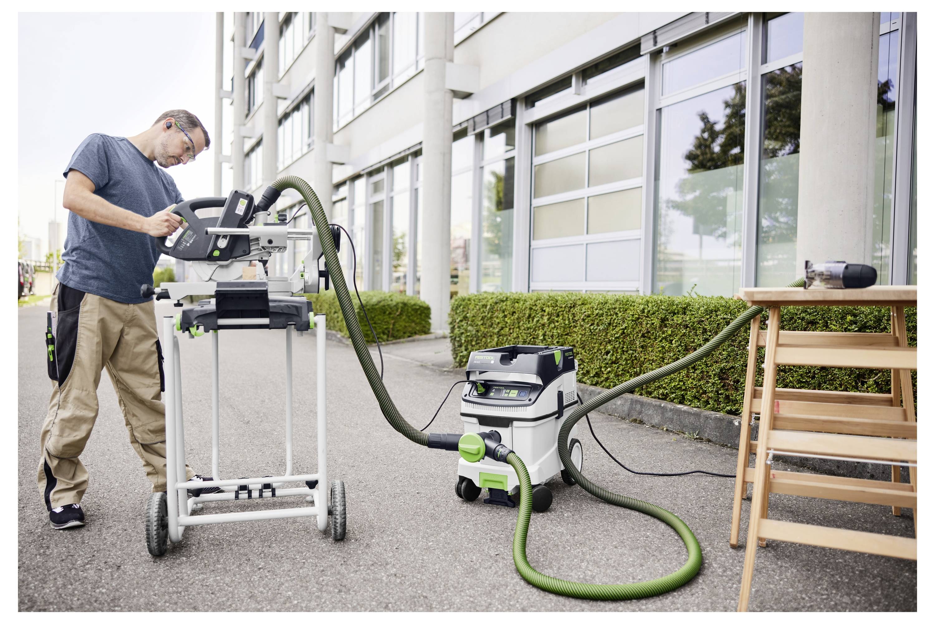 Eine Person benutzt eine Kreissäge auf einer Holzoberfläche. Die Säge ist schwarz und grün mit einem sichtbaren Blatt. Sie ist mit einem Staubsaugerschlauch verbunden.