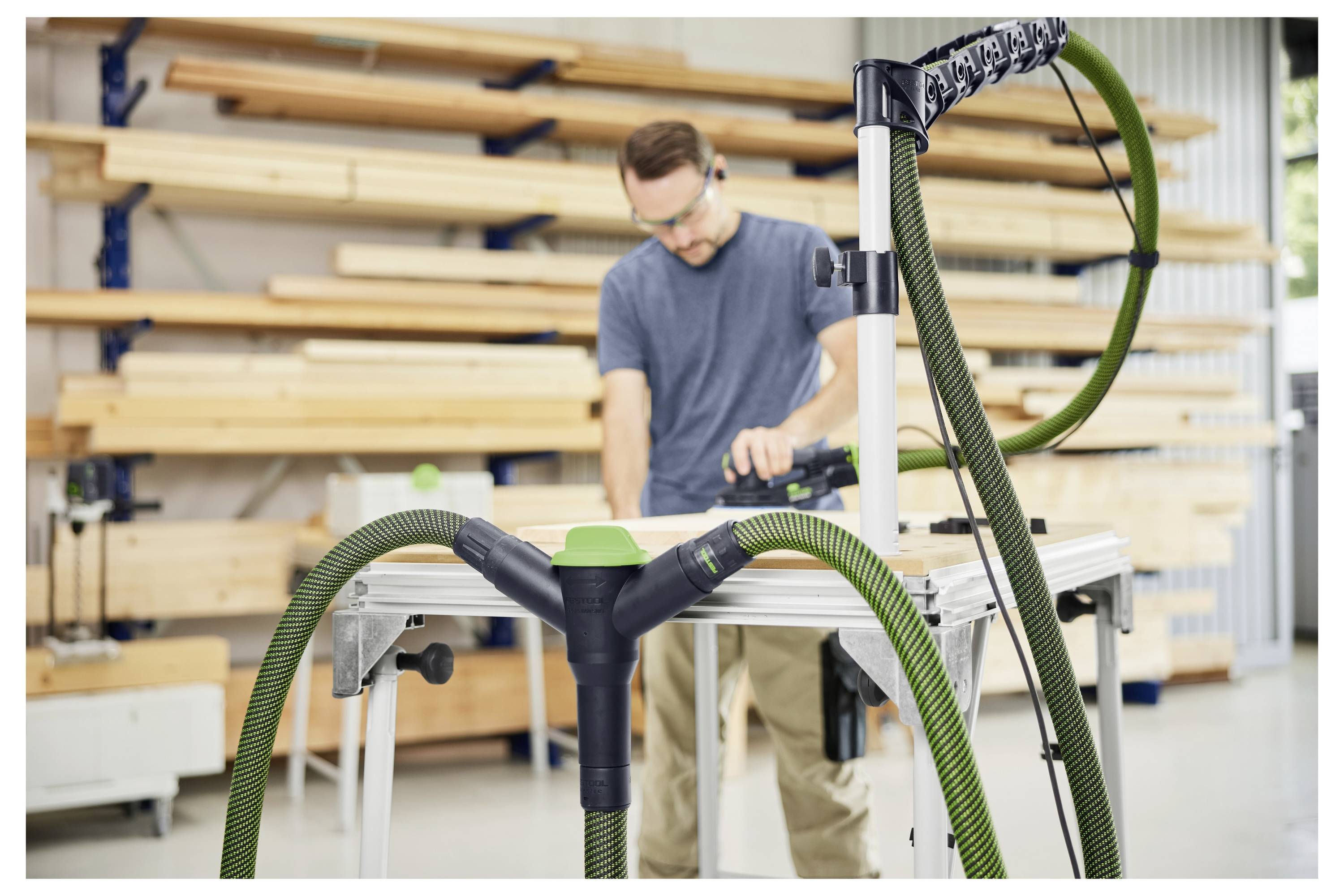 Eine Person arbeitet mit Elektrowerkzeugen an einer Werkbank in einer Holzbearbeitungswerkstatt, umgeben von Holzstapeln auf Regalen.