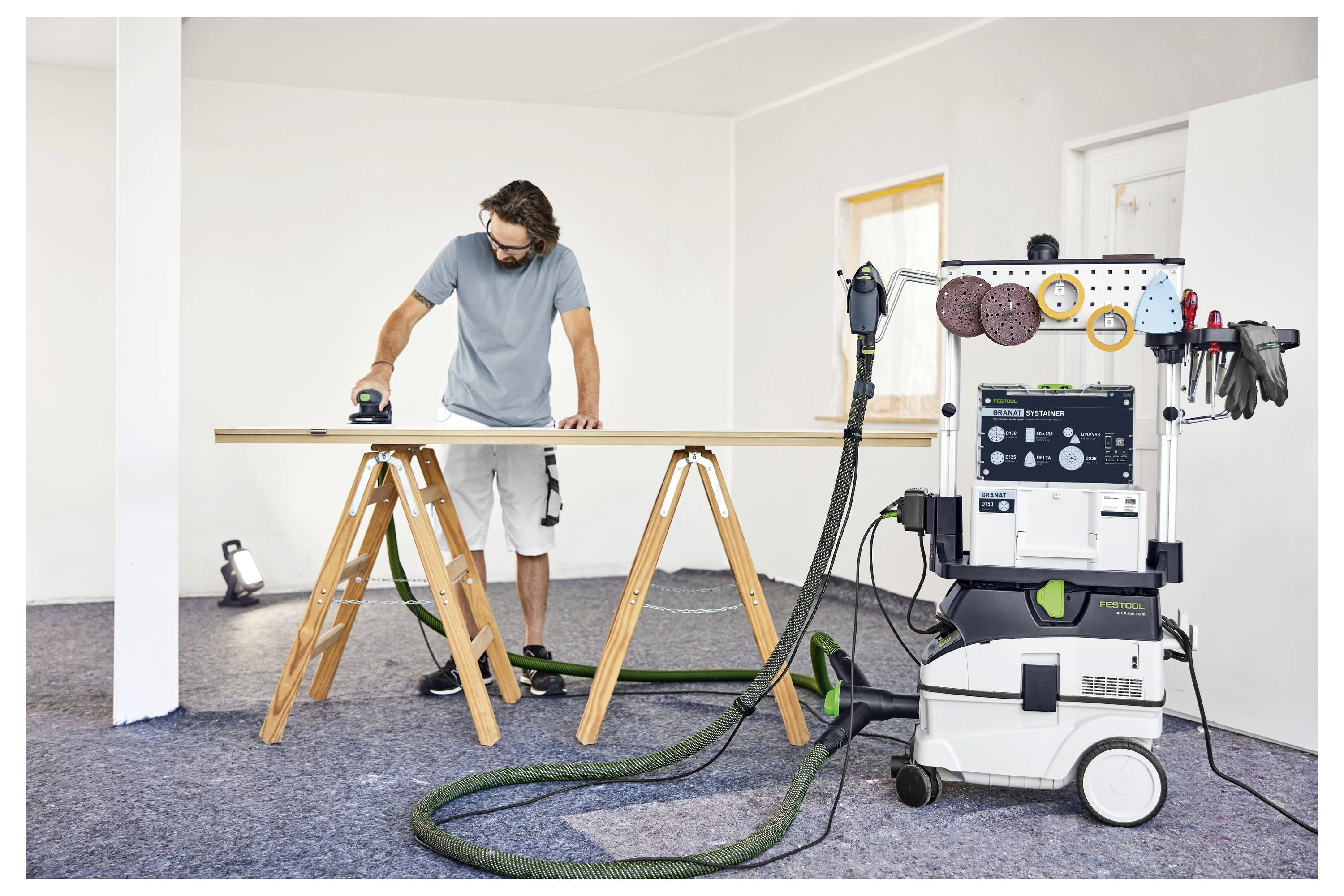 Ein Mann in einem dunklen Hemd und einer Mütze steht in einer Werkstatt und blickt auf große gestapelte Holzbretter auf Regalen.