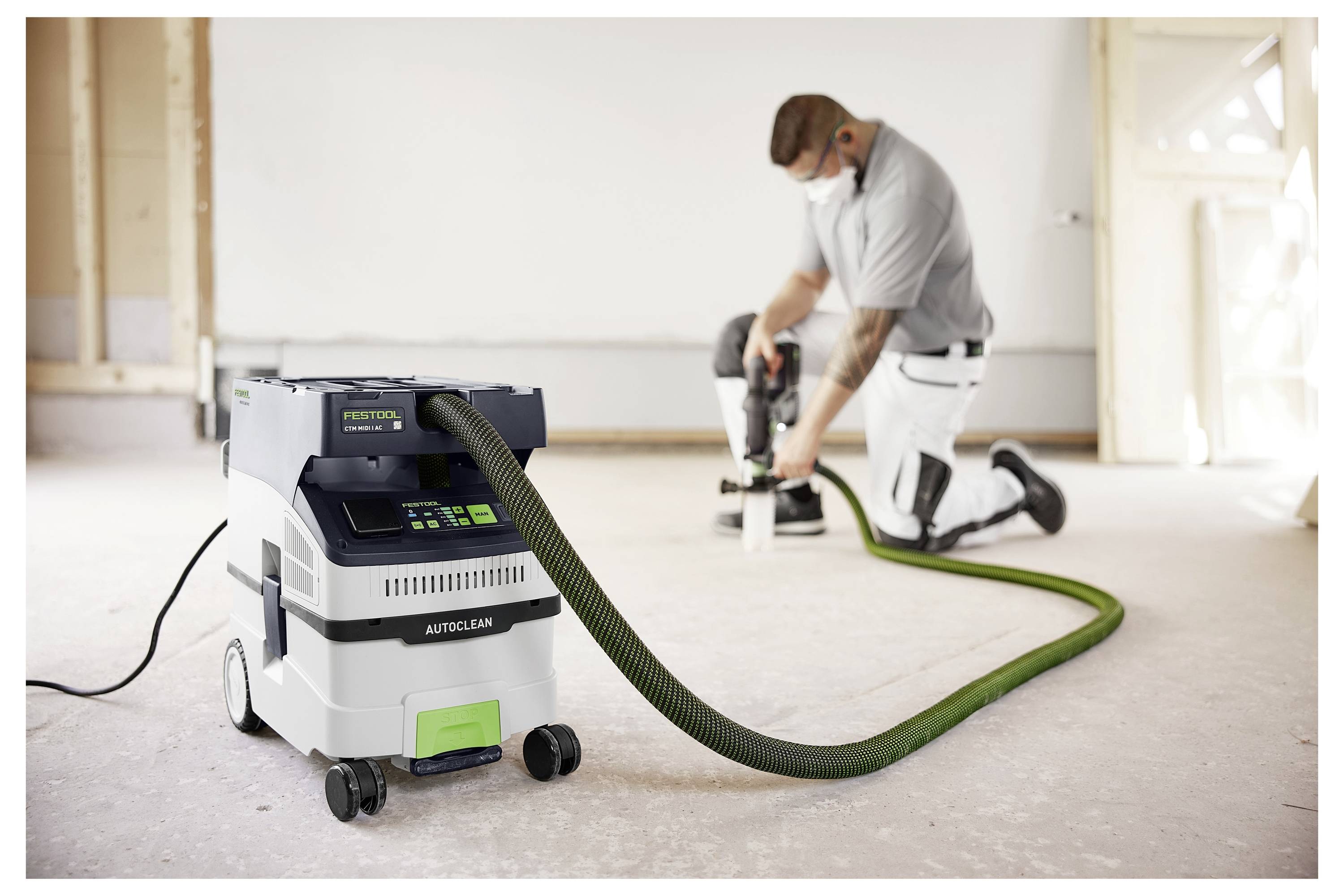 Eine Person in Arbeitskleidung hält eine Kreissäge auf einem Gerüstablage neben einem Gebäude mit vertikalen Holzleisten, was auf Bauarbeiten hindeutet.