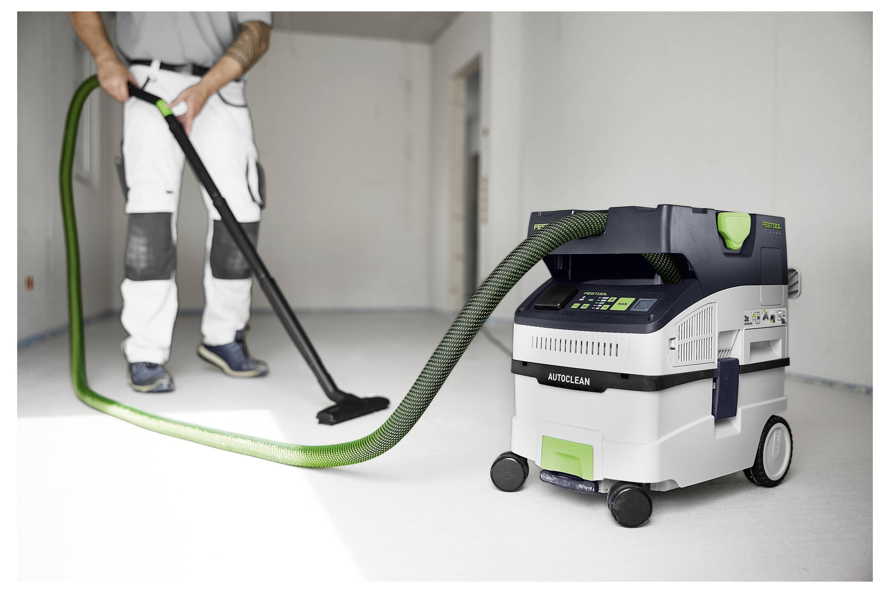 Eine Person saugt einen Raum mit einem grünen Schlauch, der an einem weißen und grünen fahrbaren Industriestaubsauger angeschlossen ist.