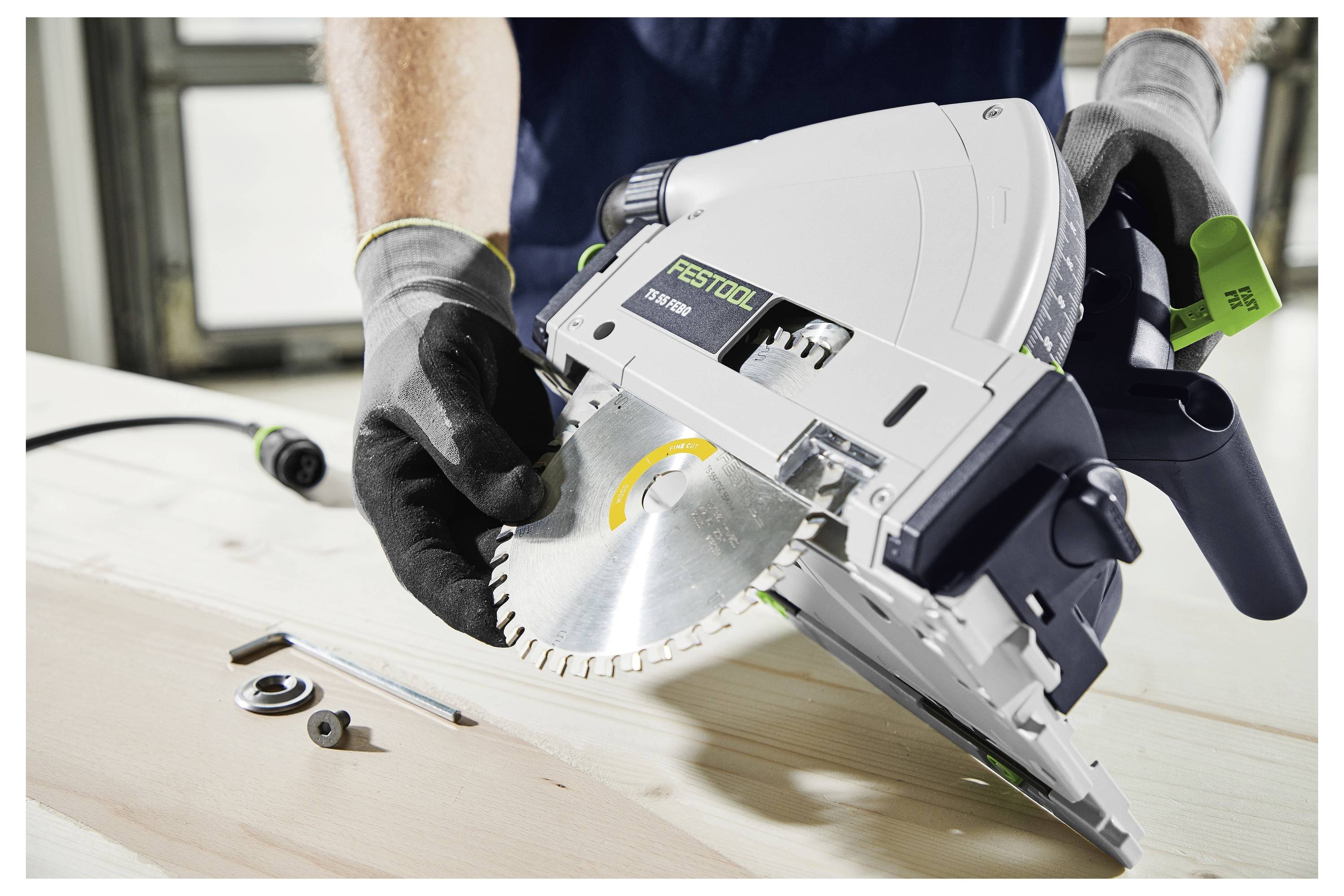 Eine Person, die Handschuhe trägt, installiert ein kreisförmiges Sägeblatt in ein Elektrowerkzeug auf einer Werkbank, mit Werkzeugen und Teilen in der Nähe.