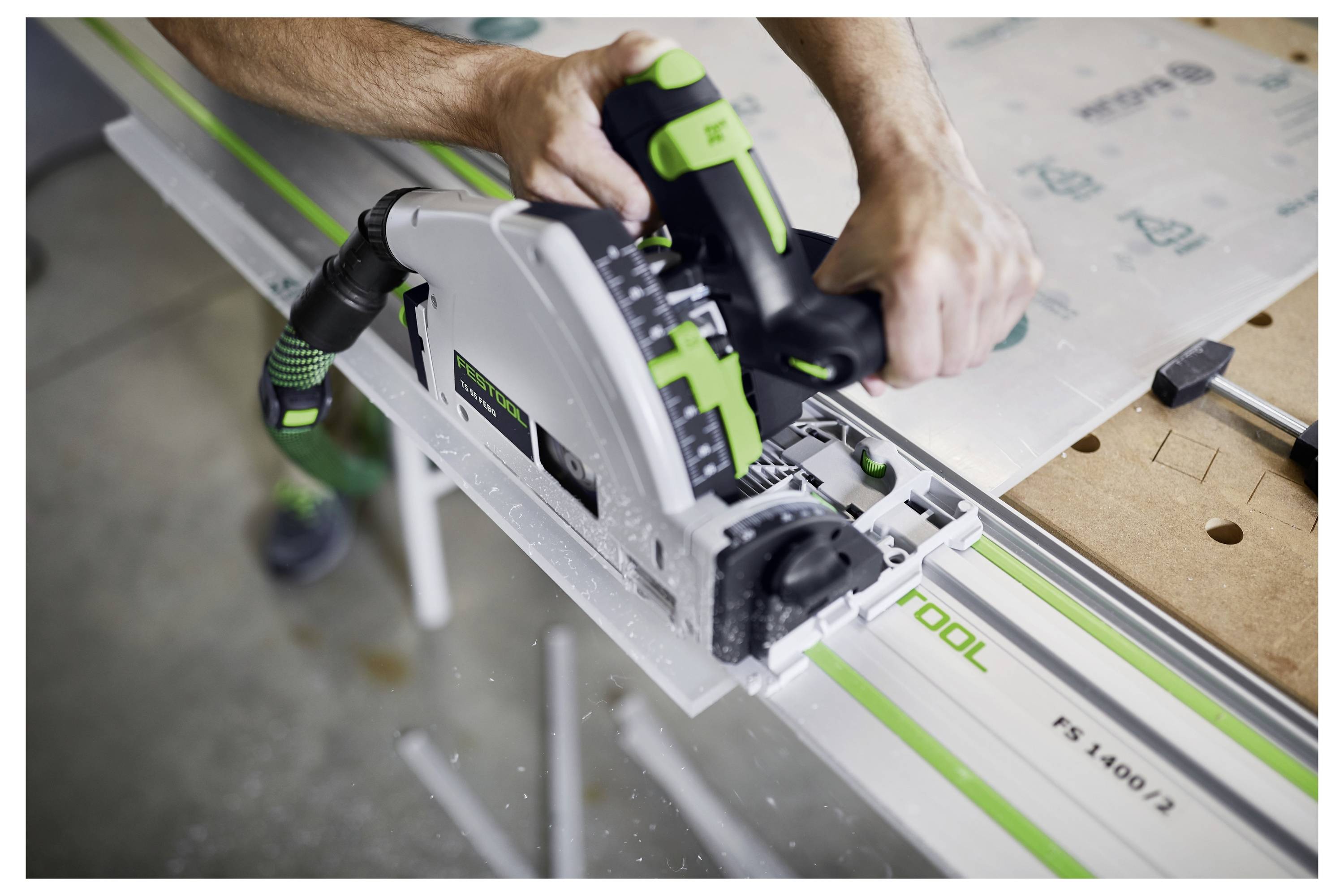 Eine Person benutzt eine Kreissäge, um entlang einer Führungsschiene auf einer Werkbank zu schneiden. Die Säge hat eine grün-weiße Farbgebung.