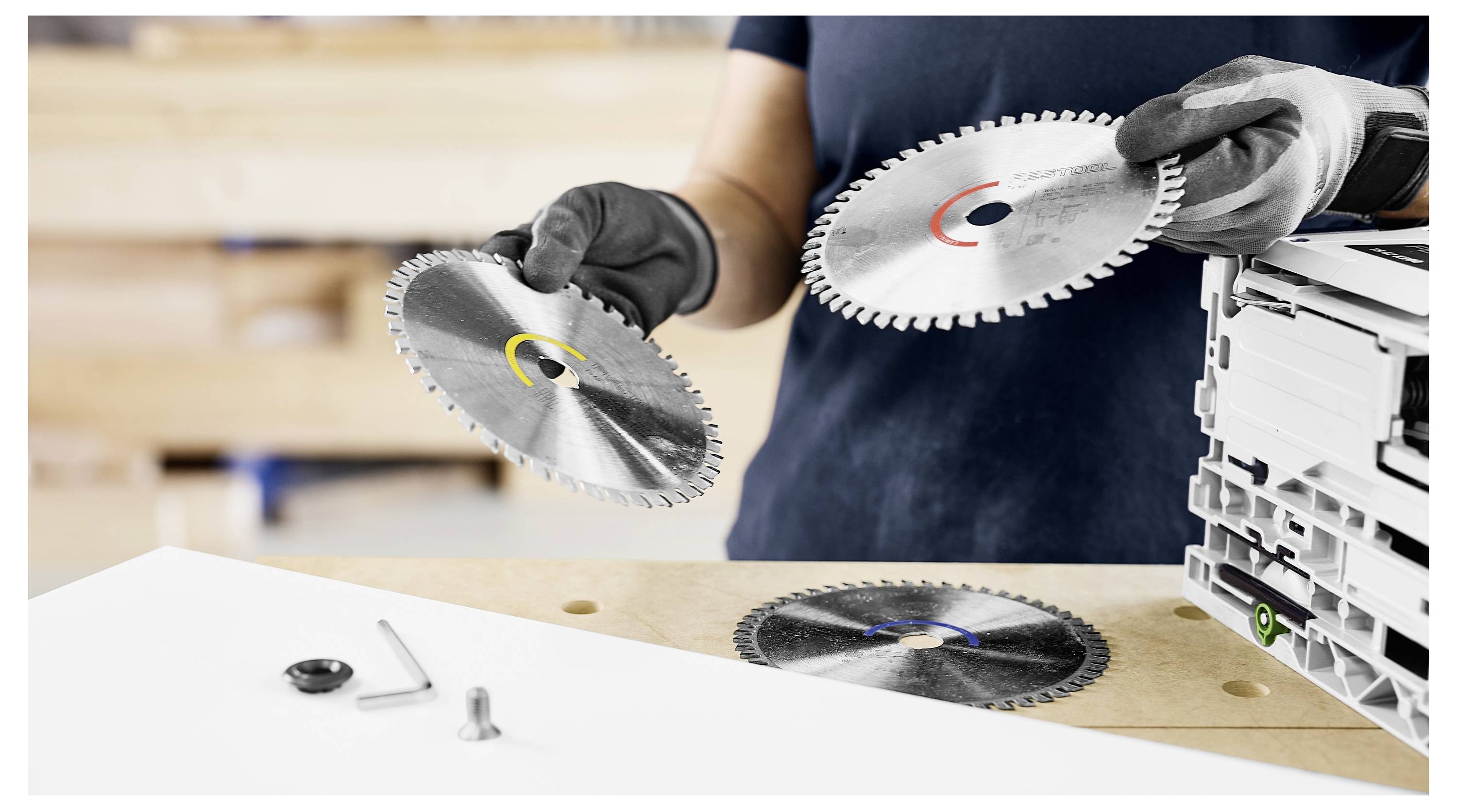 Eine Person mit Handschuhen hält zwei Kreissägeblätter, eines in jeder Hand, über einer Werkbank. Ein weiteres Blatt und ein paar Werkzeuge liegen auf der Bank.