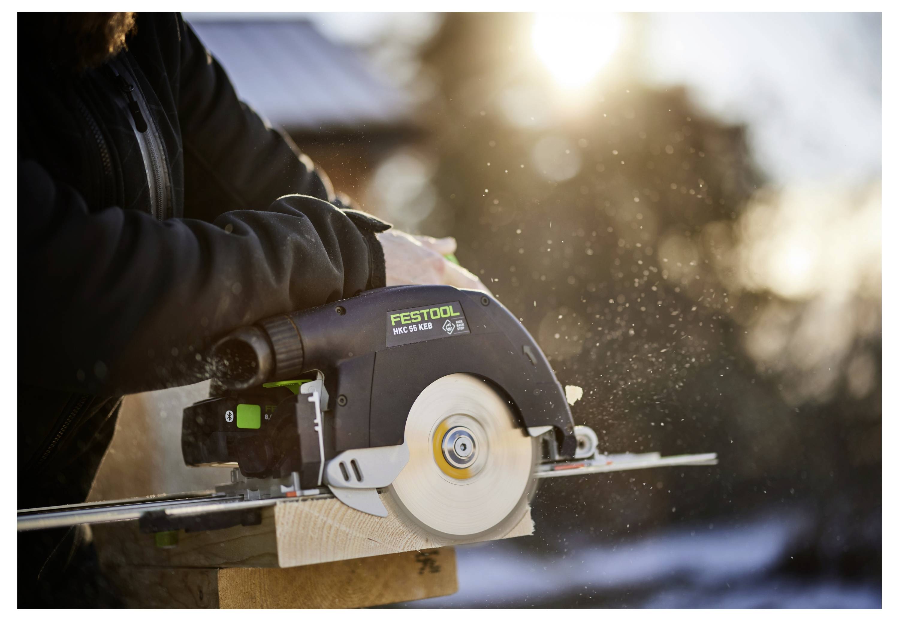Eine Person benutzt eine Kreissäge, um ein Holzbrett im Freien bei Sonnenuntergang zu schneiden. Staubpartikel sind in der Luft sichtbar, mit sanftem Sonnenlicht im Hintergrund.
