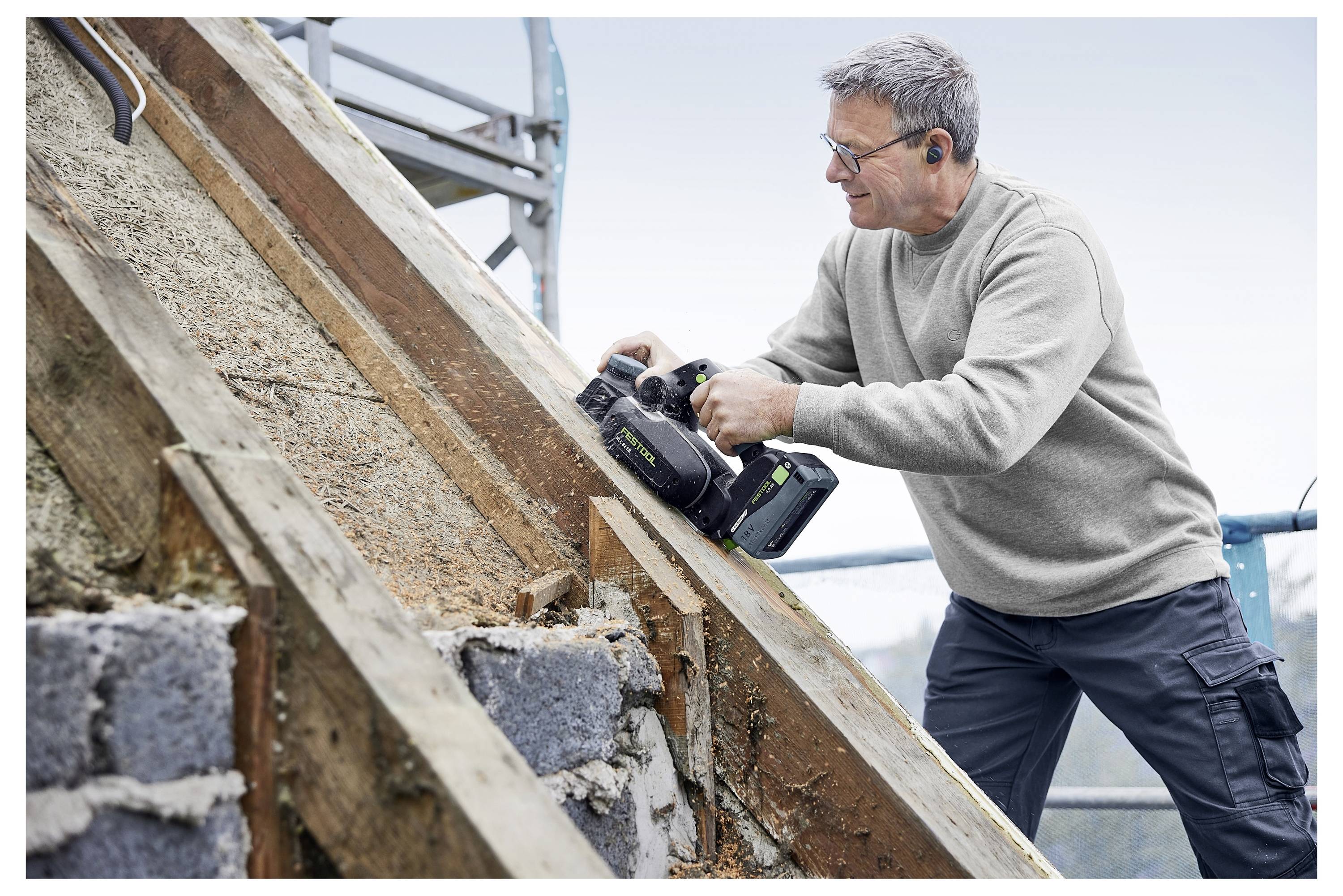 Ein Mann in einem Pullover benutzt ein Elektrowerkzeug an einem Holzrahmen auf einer Baustelle und trägt dabei Schutzbrille und Gehörschutz.