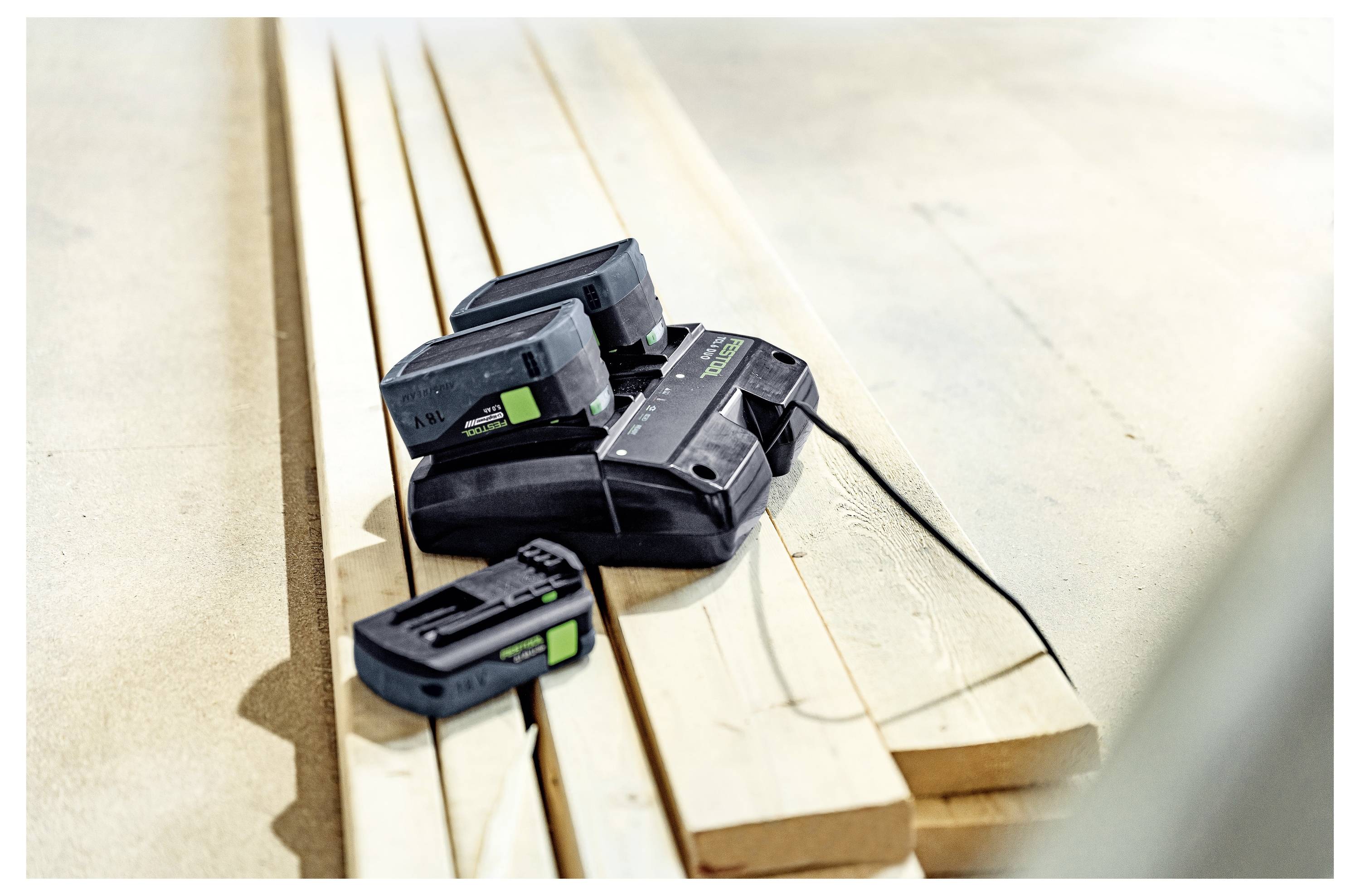 Ein Satz von vier Akkupacks für Elektrowerkzeuge auf einem Ladegerät, platziert auf Holzbrettern, was auf eine Werkstatt- oder Baustelle hindeutet.
