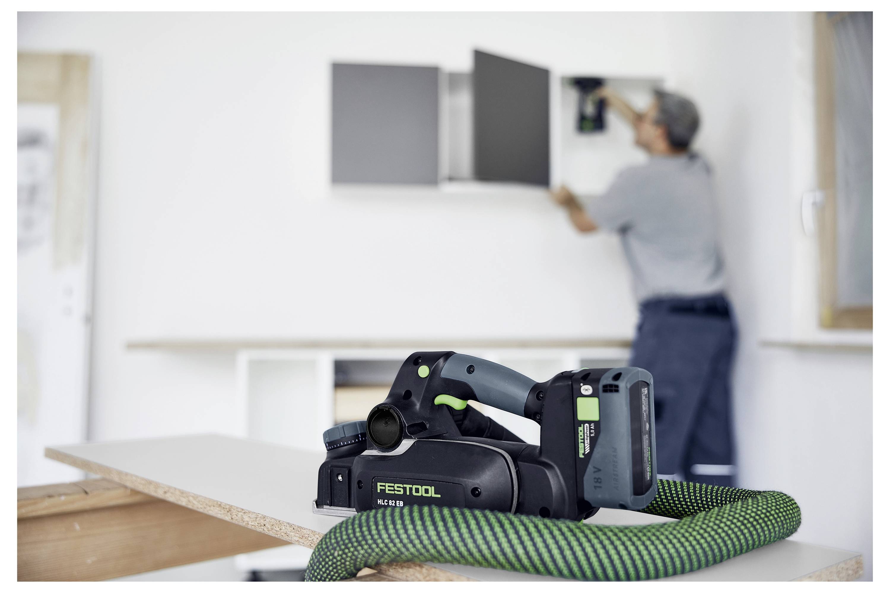 Ein tragbares Festool-Werkzeug im Fokus auf einem Tisch, mit einem Arbeiter, der im unscharfen Hintergrund einen Schrank installiert, was auf ein Baustellen-Setting hindeutet.