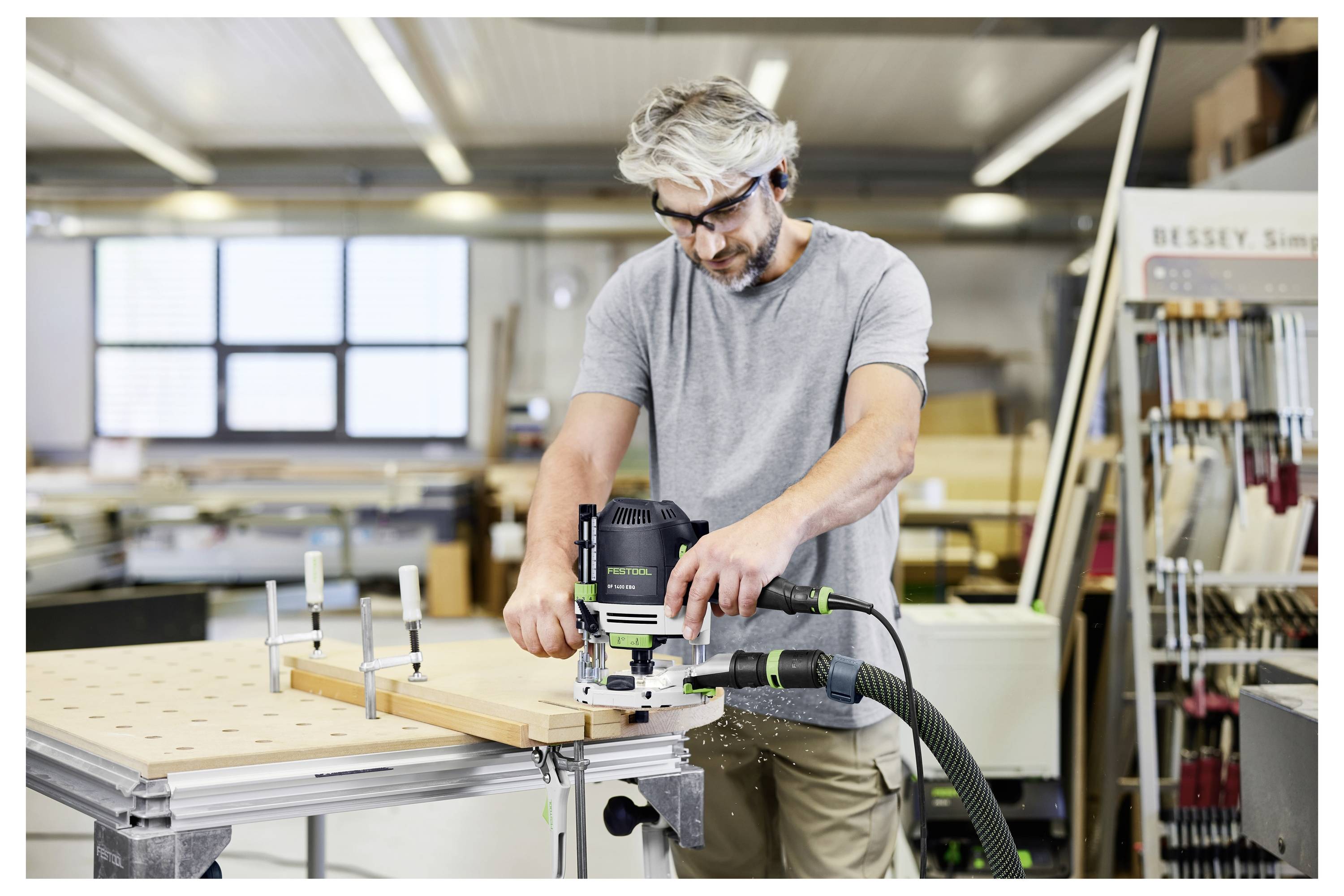 Eine Person mit grauem Haar bedient ein Elektrowerkzeug an einem Holzstück in einer Werkstatt und trägt Schutzausrüstung. Werkzeuge und Materialien befinden sich im Hintergrund.
