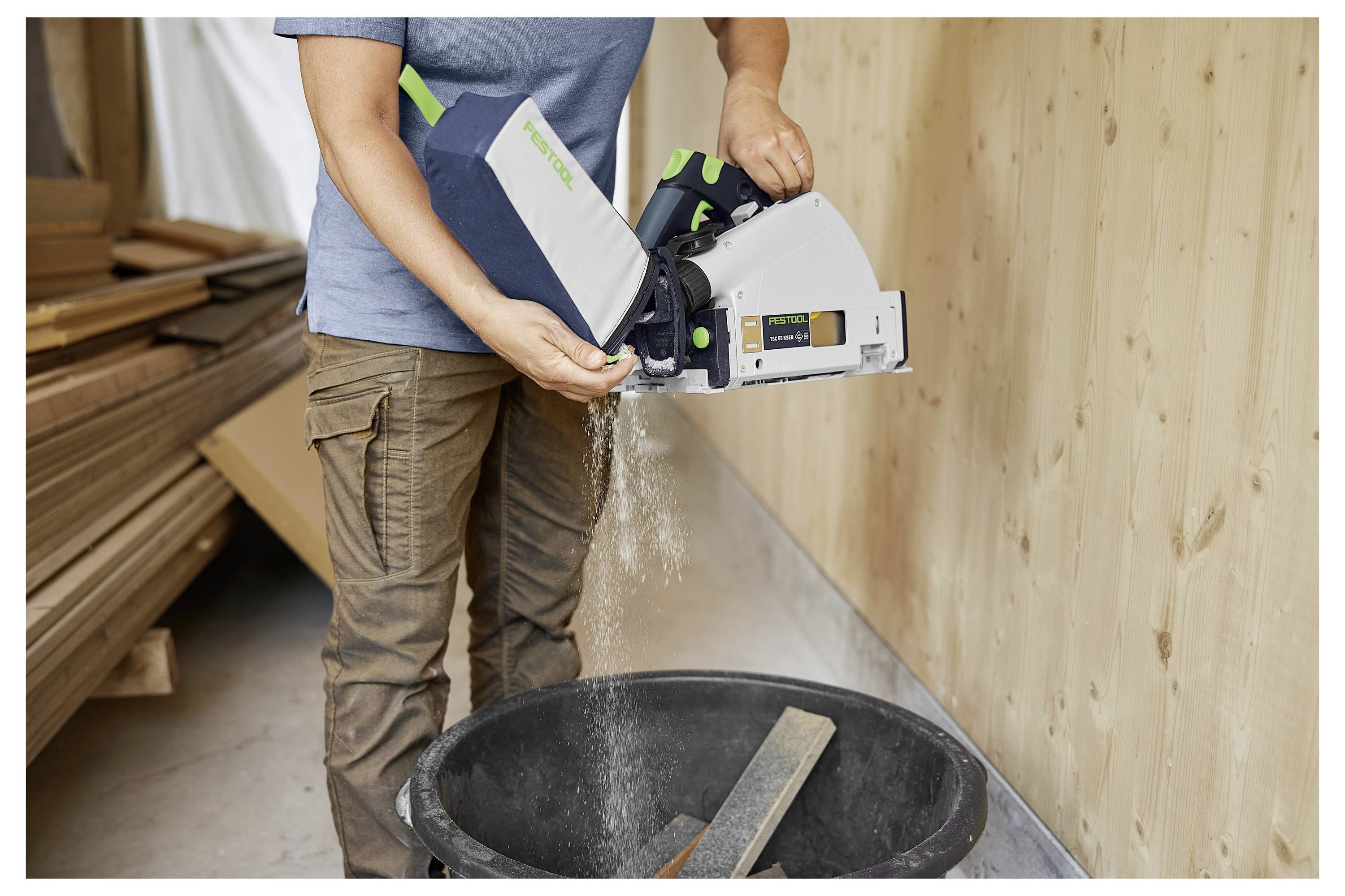 Eine Person leert Holzspäne aus einem Staubsammler in einer Werkstatt. Holzstapel lehnen sich in der Nähe an eine Wand.