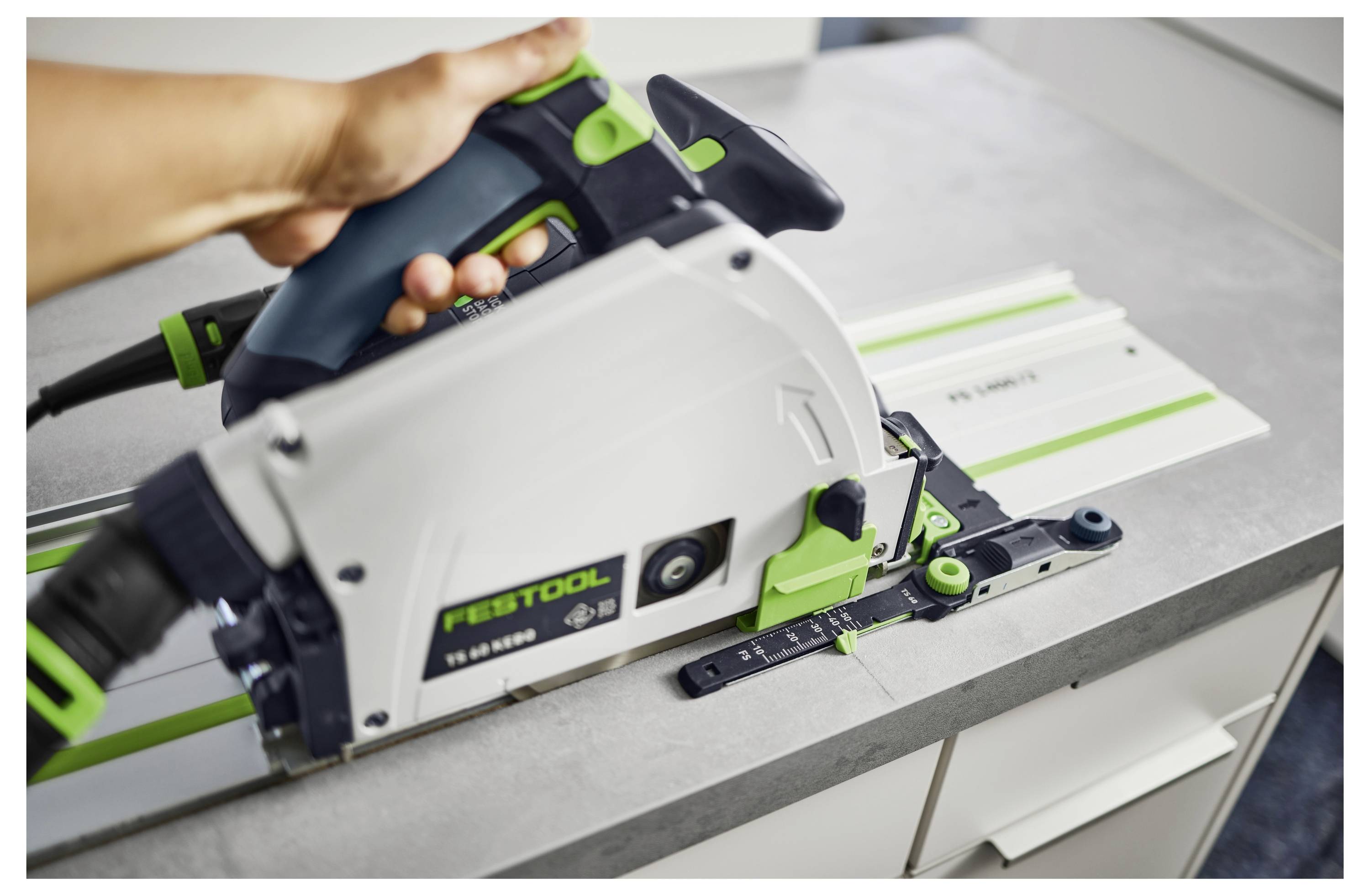 Eine Hand bedient eine Festool-Handkreissäge auf einer grauen Arbeitsfläche und führt die Säge entlang einer Metallschiene für präzises Schneiden.