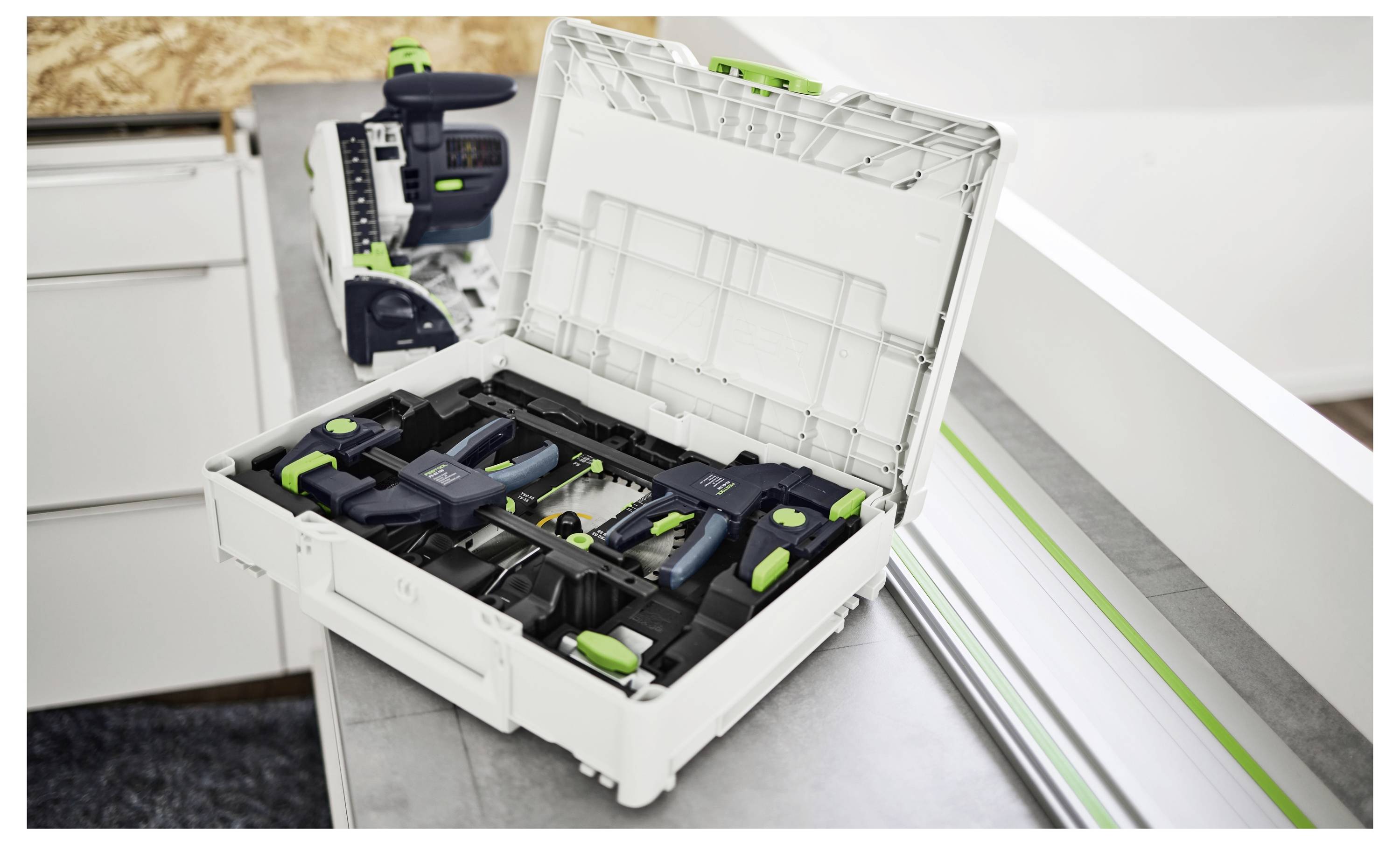 Offene weiße Werkzeugkiste auf einer Werkbank mit verschiedenen grünen und schwarzen Werkzeugen und einer teilweise sichtbaren Maschine im Hintergrund.